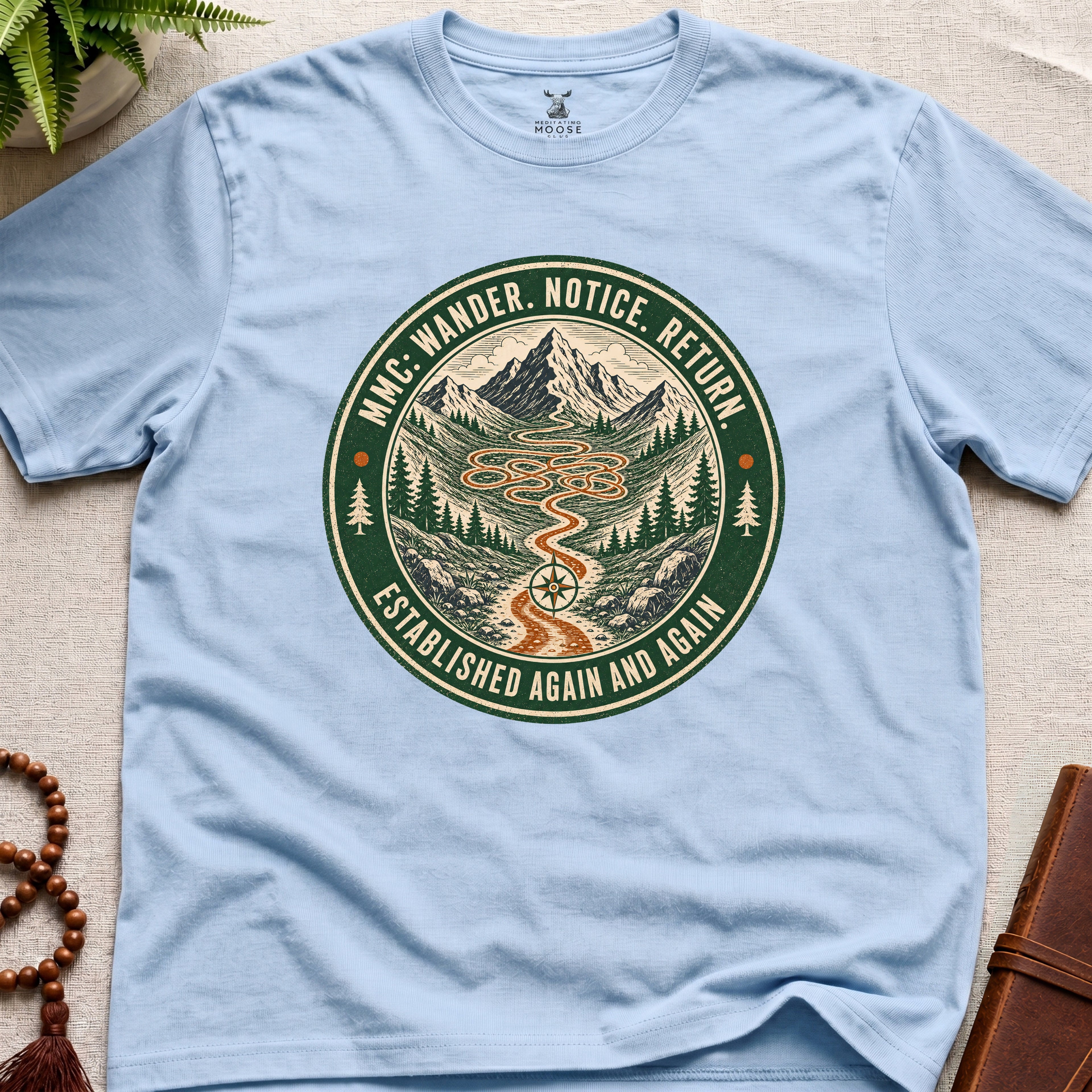 Light blue mindfulness t-shirt mockup featuring Meditating Moose Club Wander Notice Return mountain trail badge graphic