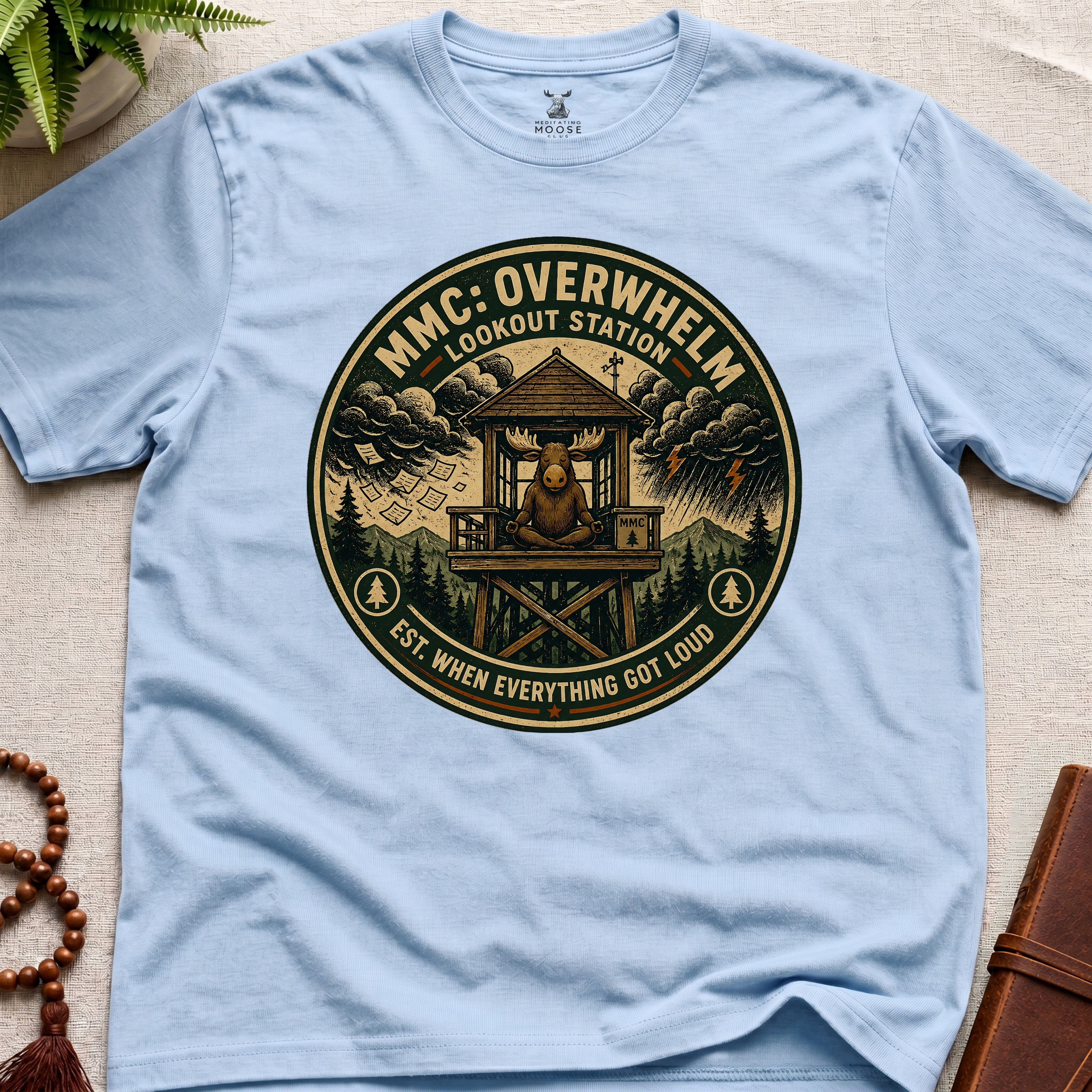 Light blue mindfulness t-shirt mockup featuring Overwhelm Lookout Station meditating moose stress graphic