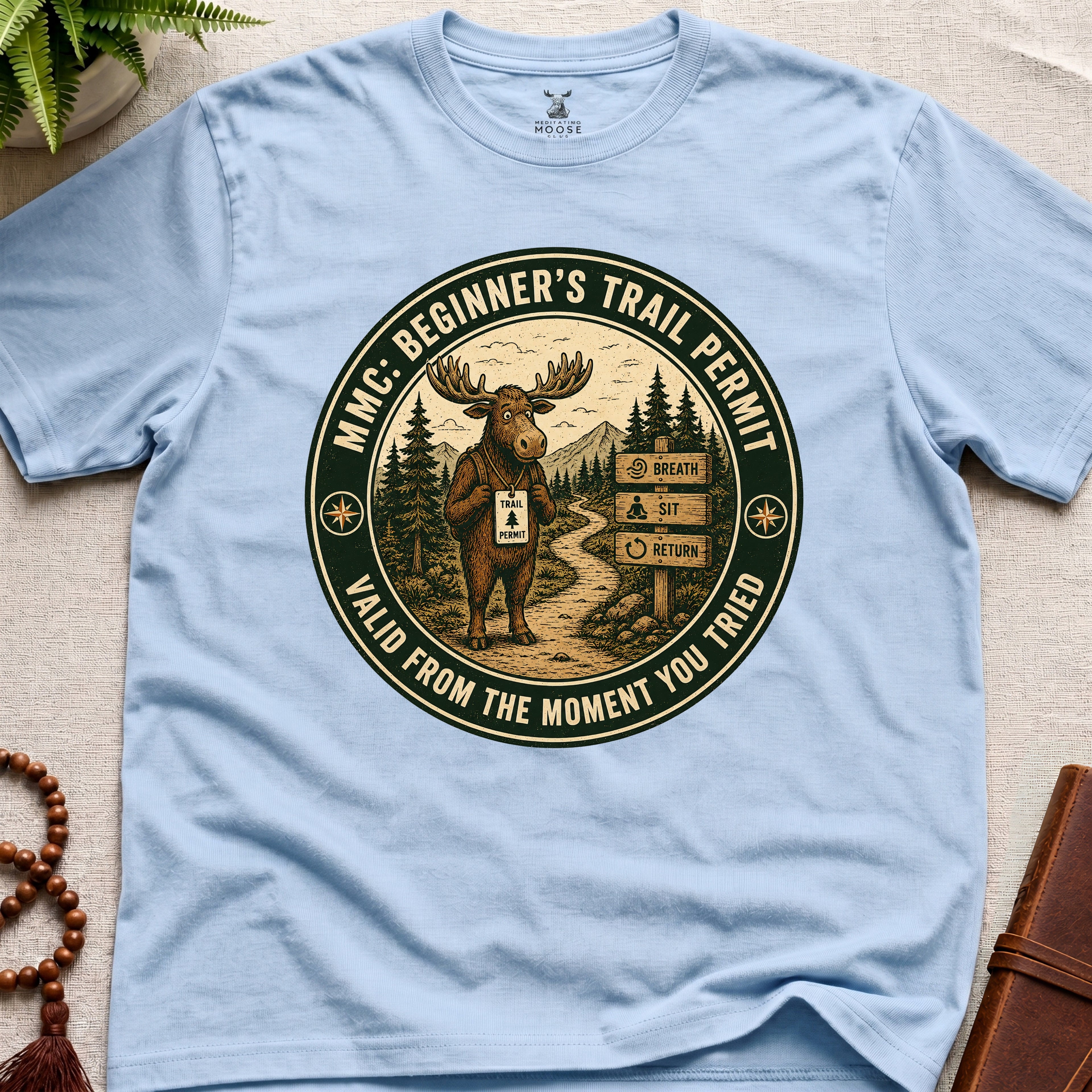 Light blue mindfulness t-shirt mockup featuring Beginner’s Trail Permit meditating moose beginner meditation graphic