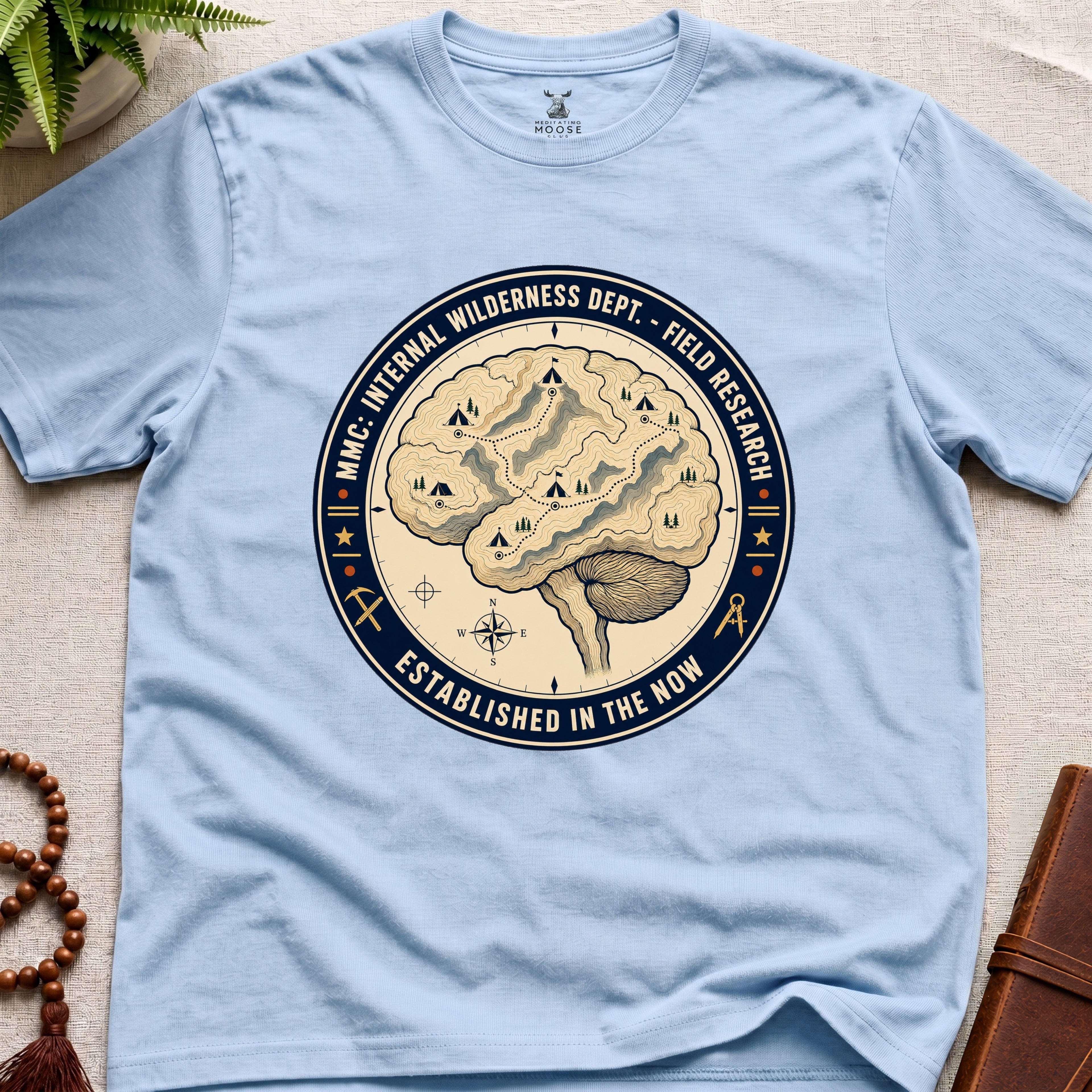 Light blue t-shirt with a circular graphic design featuring a brain and wilderness elements on a beige surface.
