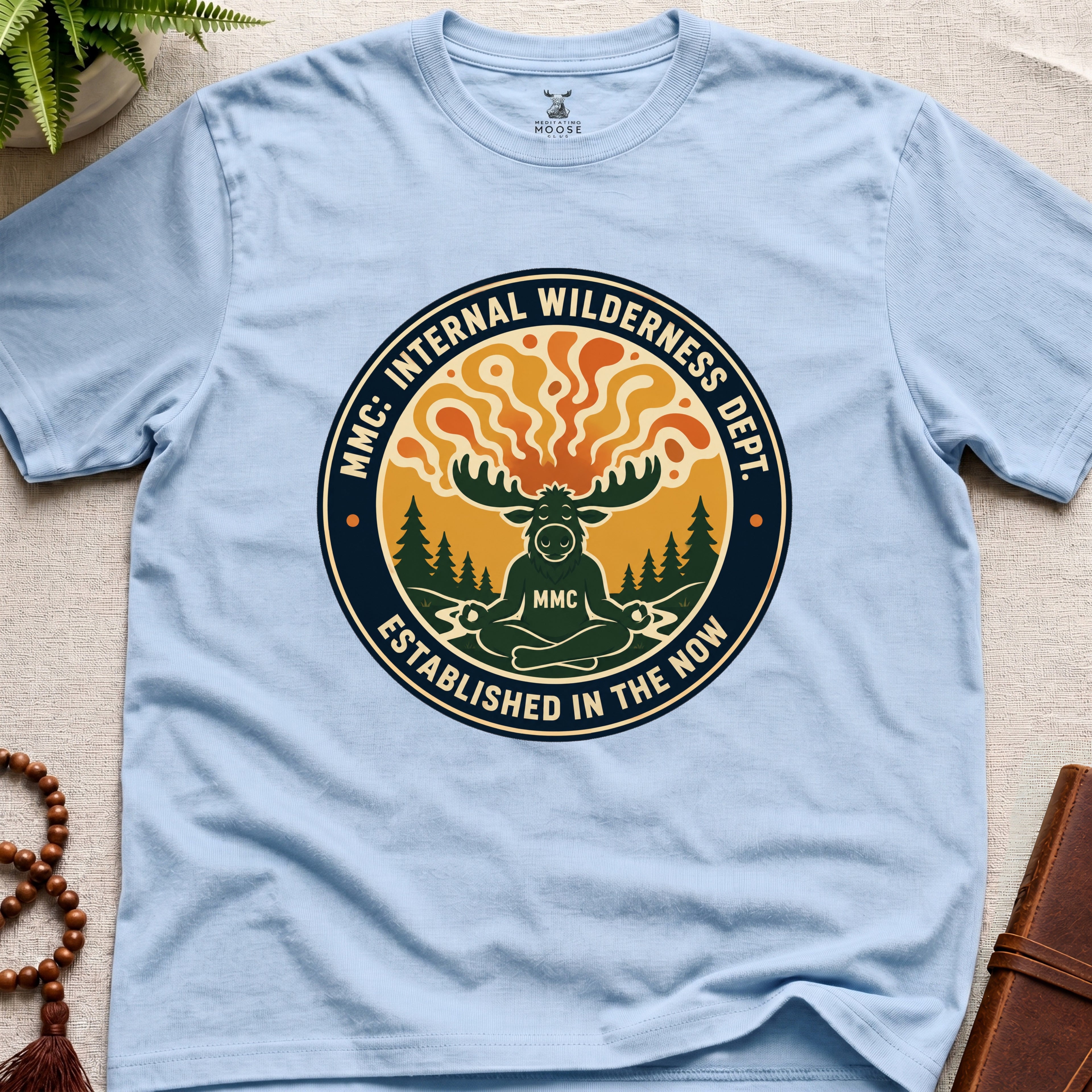Light blue t-shirt with a circular graphic design featuring a moose and trees on a beige surface.