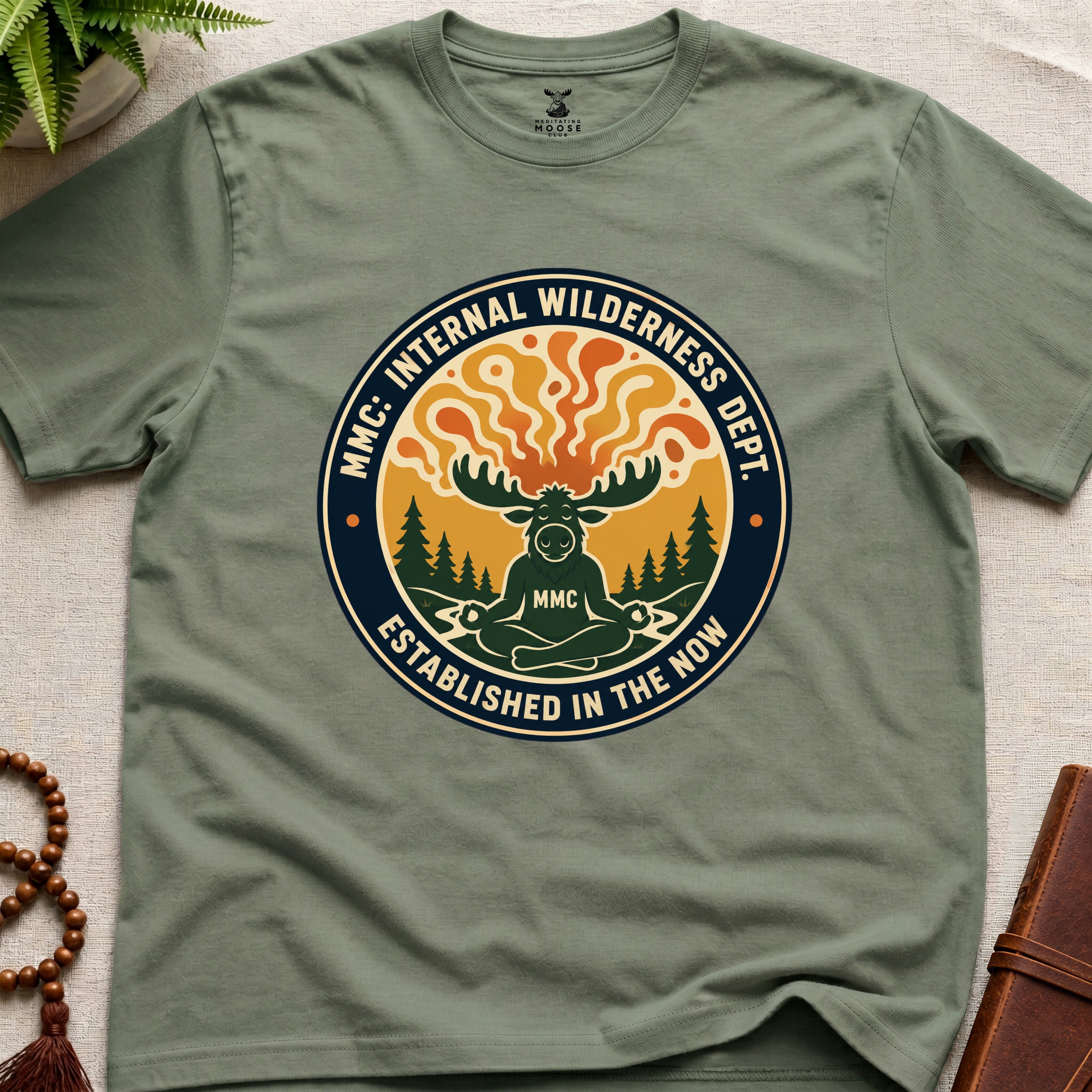 Military green t-shirt with a circular graphic design featuring a moose and trees on a beige surface.