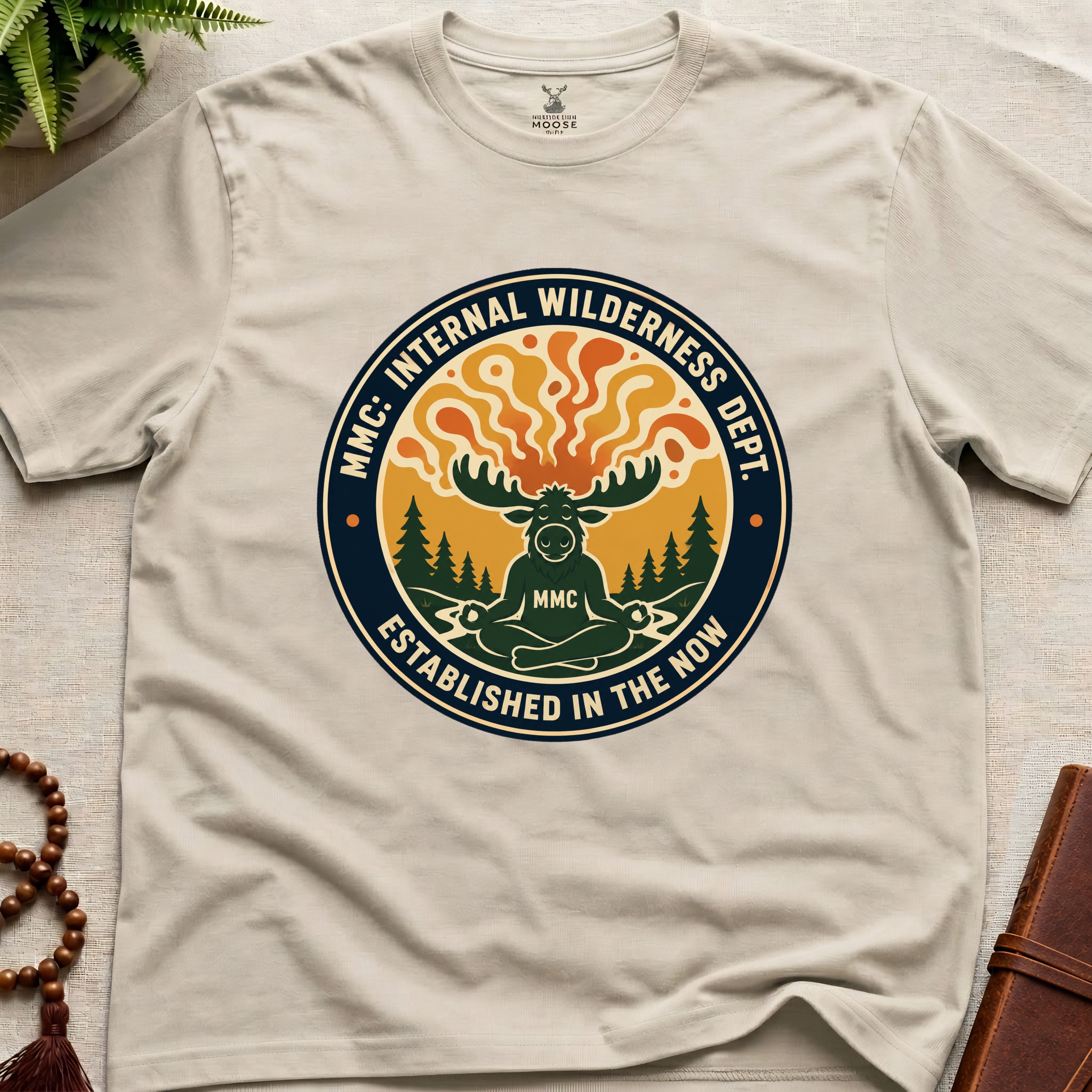 Beige t-shirt with a circular graphic design featuring a moose and trees on a light wooden surface.