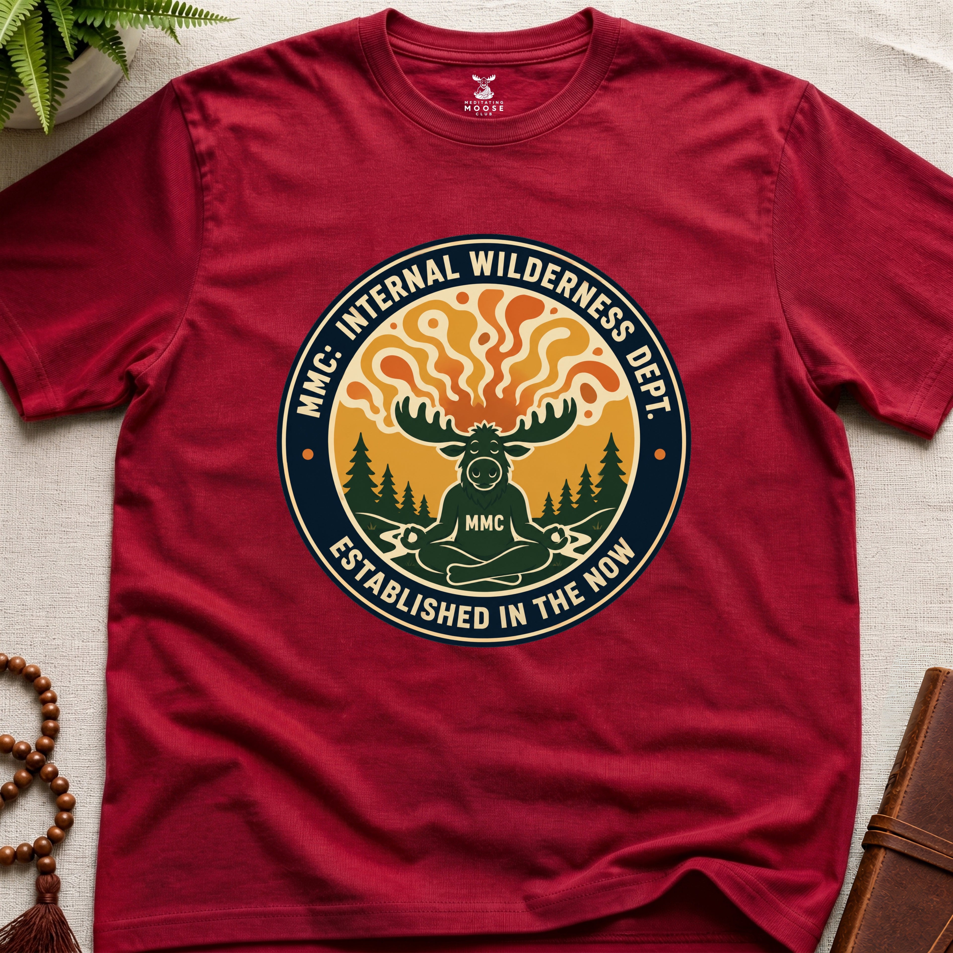 Red t-shirt with a circular graphic design featuring a moose and trees on a beige surface.