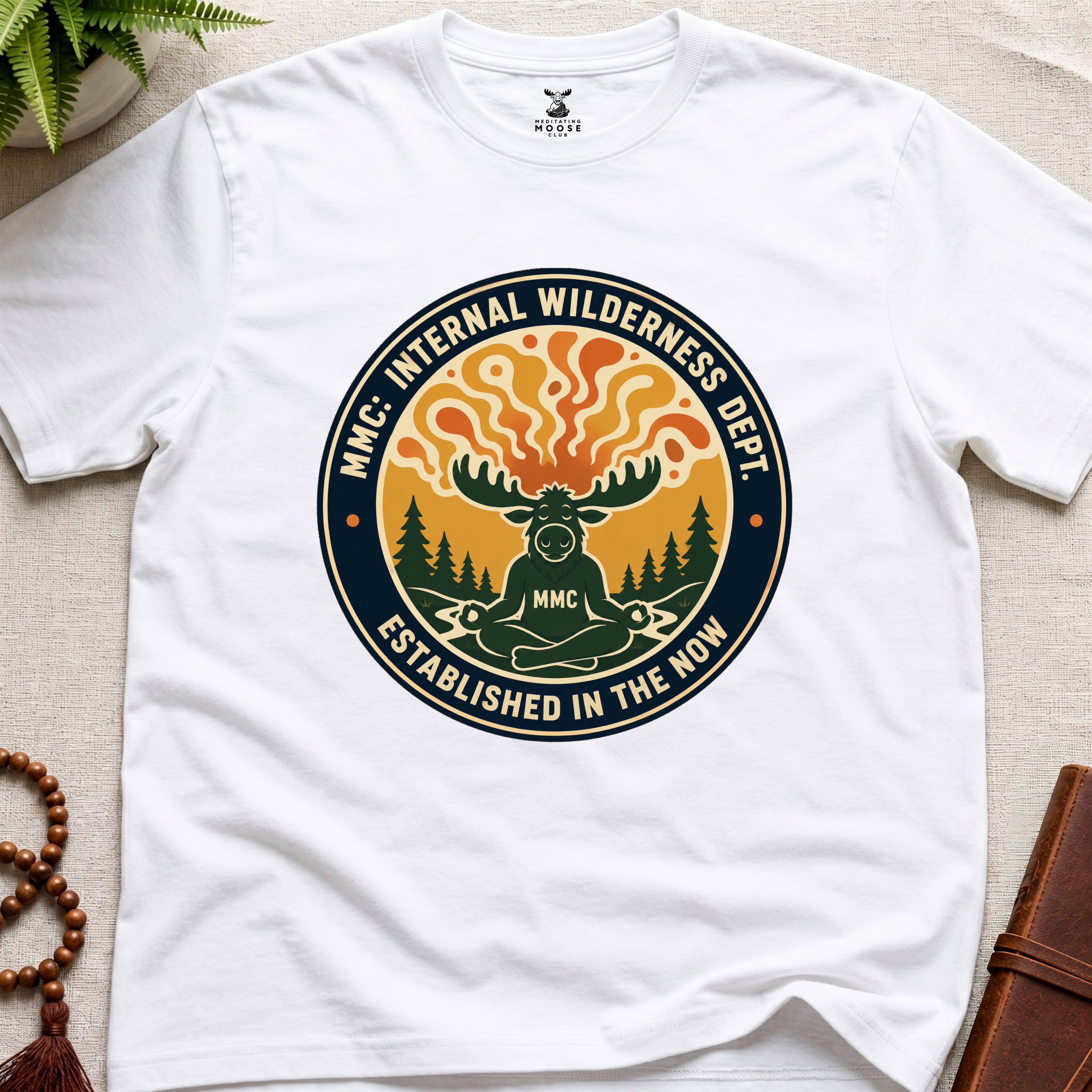 White t-shirt with a circular graphic design featuring a moose and trees on a beige surface.