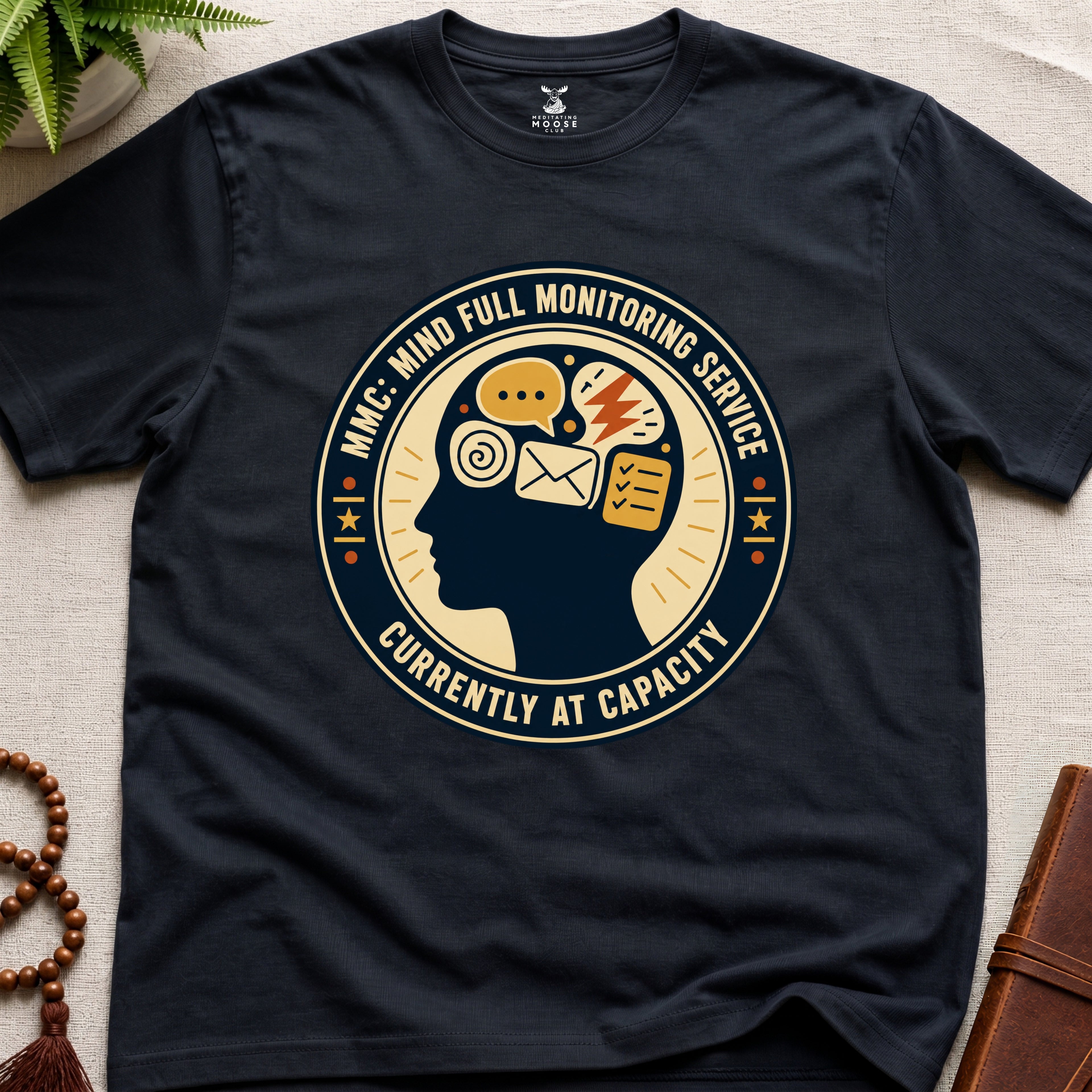 Black t-shirt with a badge graphic design featuring a head full of icons and text "Mind Full Monitoring Service" on a beige surface