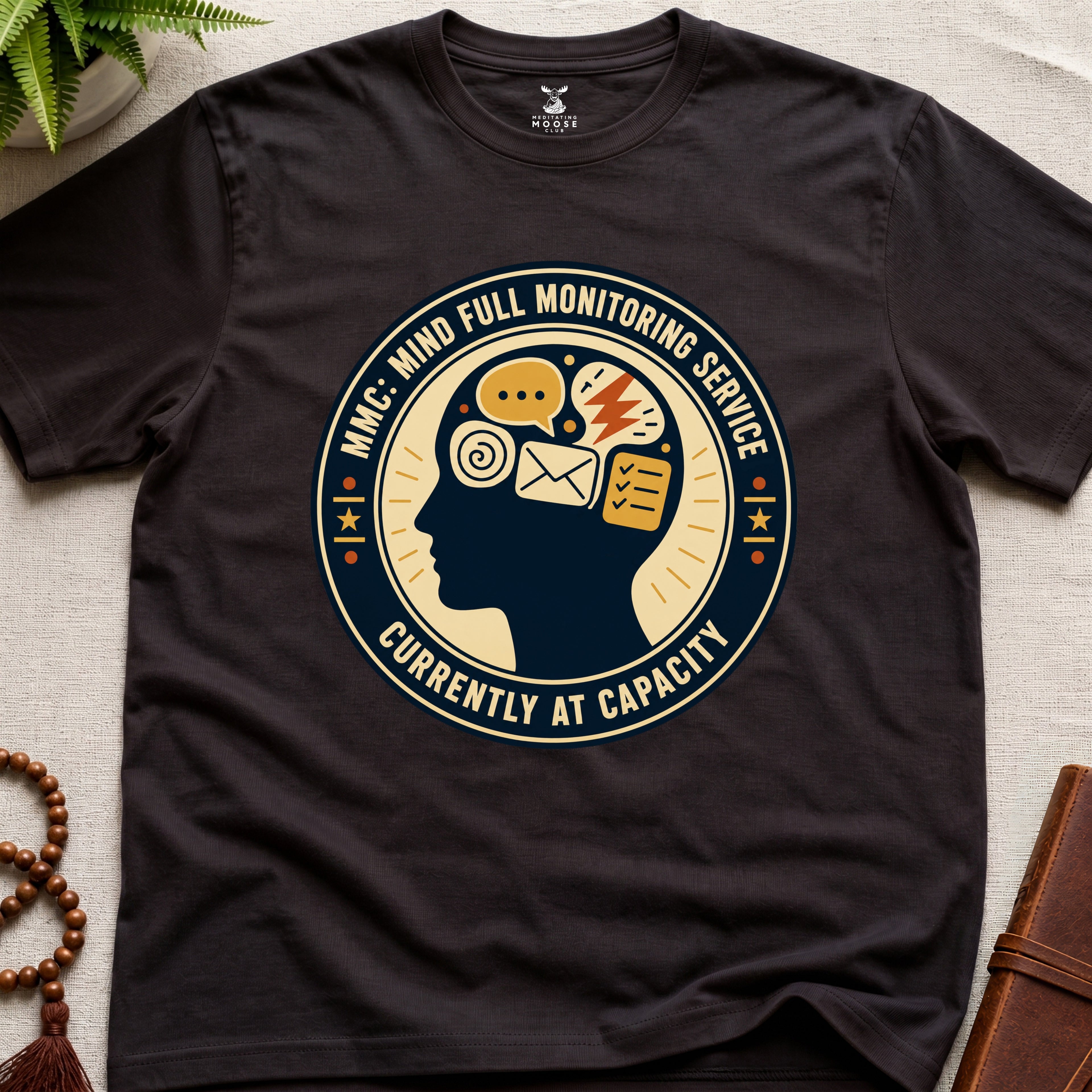 Dark brown t-shirt with a badge graphic design featuring a head full of icons and text "Mind Full Monitoring Service" on a beige surface