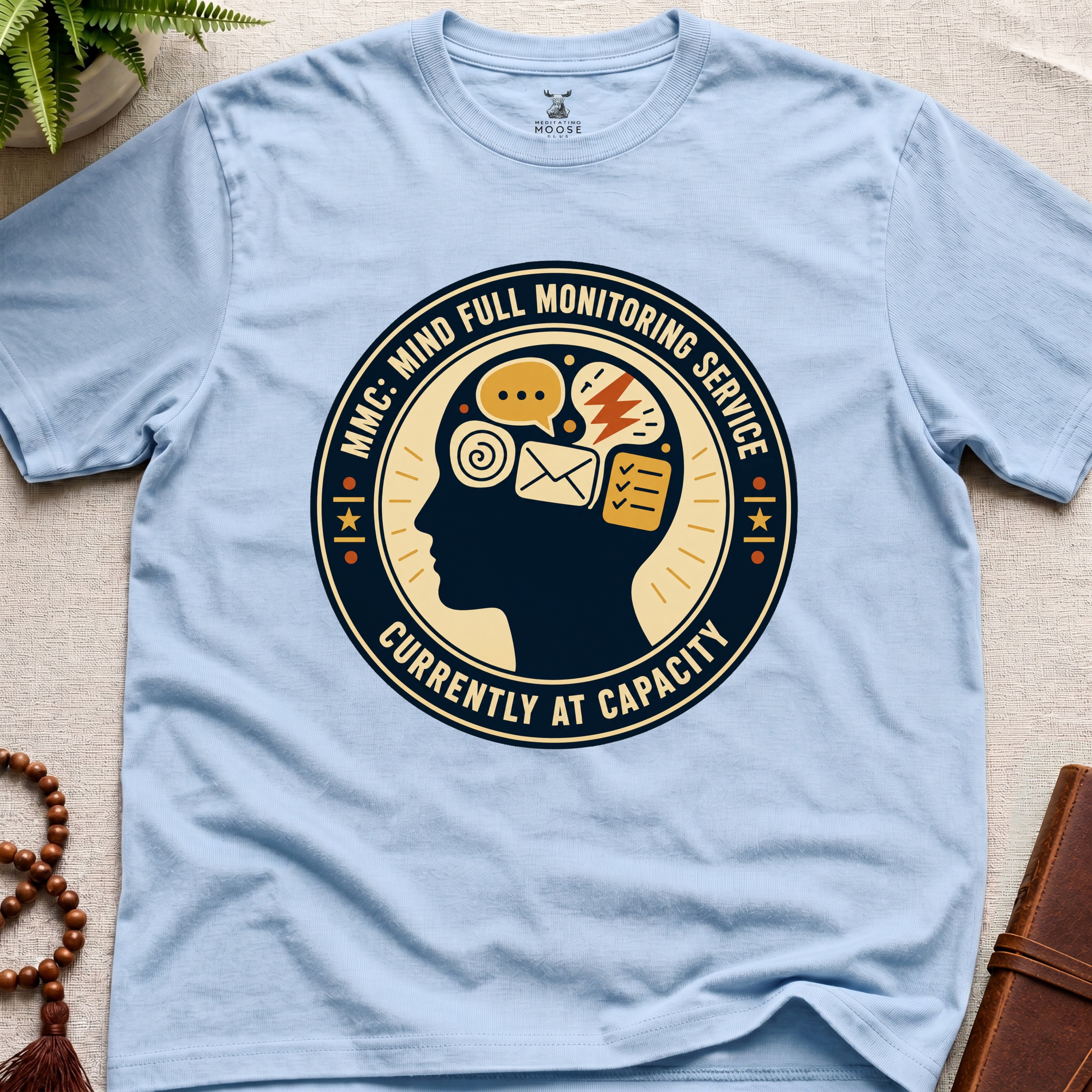 Light blue t-shirt with a badge graphic design featuring a head full of icons and text "Mind Full Monitoring Service" on a beige surface