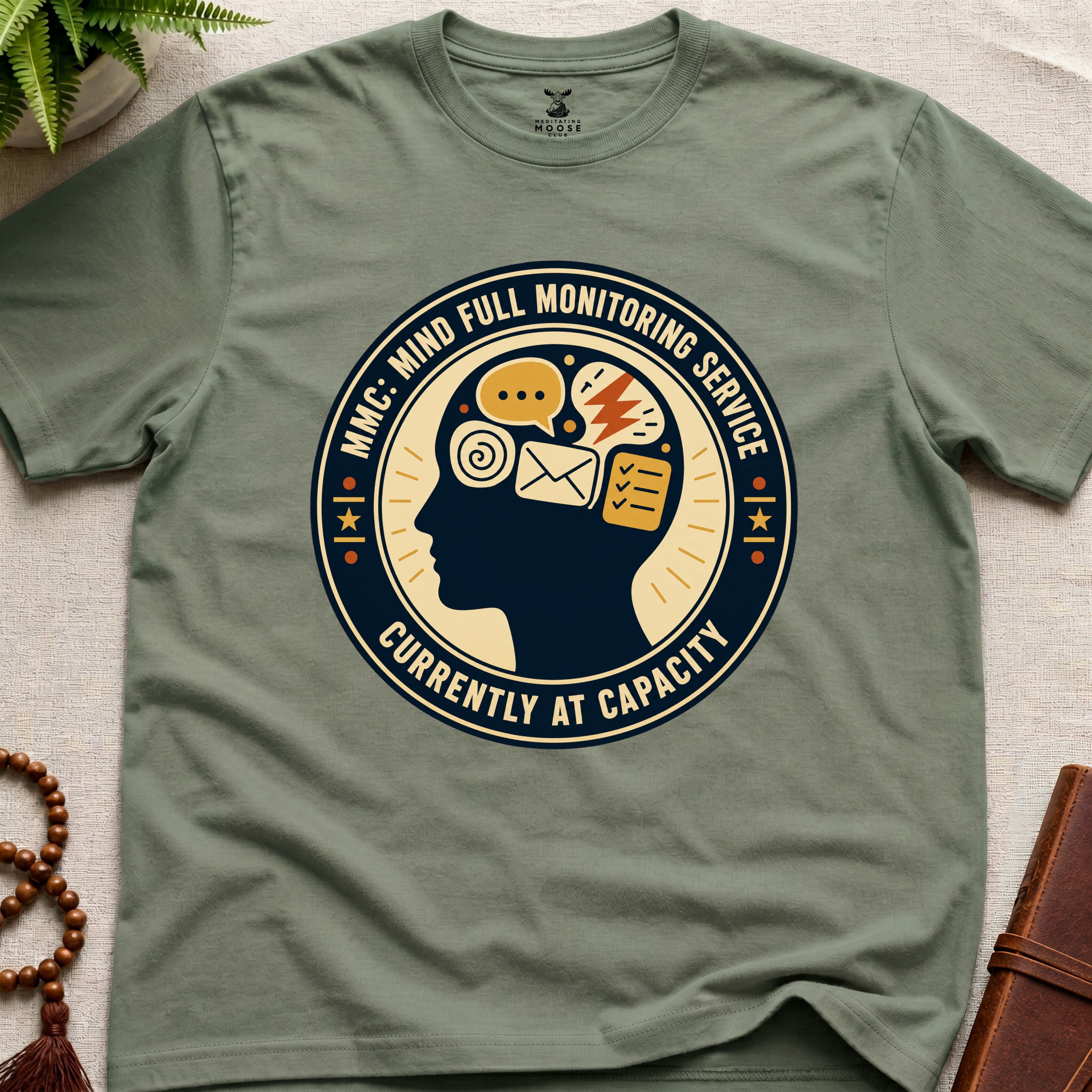 Military green t-shirt with a badge graphic design featuring a head full of icons and text "Mind Full Monitoring Service" on a beige surface