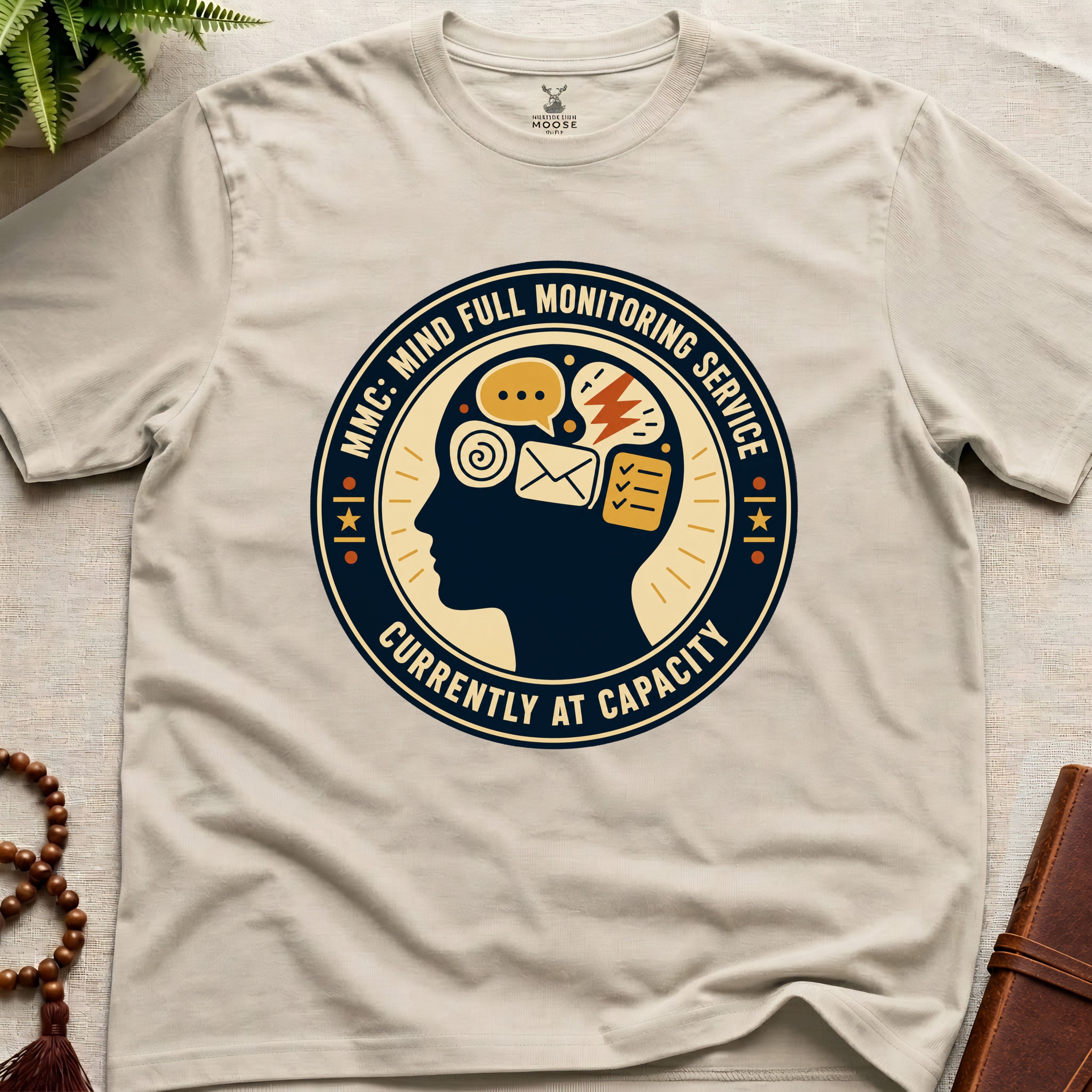 Beige t-shirt with a badge graphic design featuring a head full of icons and text "Mind Full Monitoring Service" on a beige surface