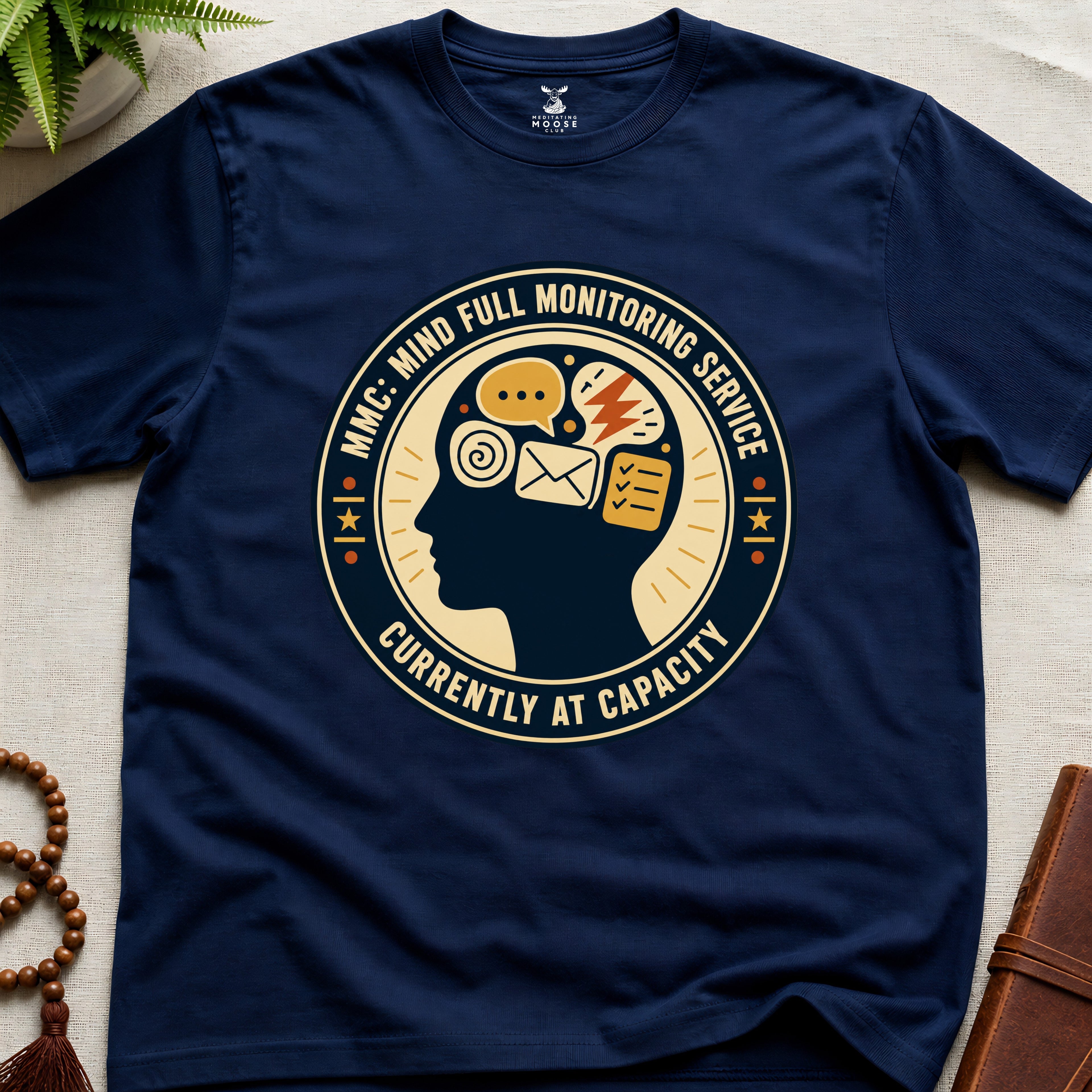 Navy blue t-shirt with a badge graphic design featuring a head full of icons and text "Mind Full Monitoring Service" on a beige surface