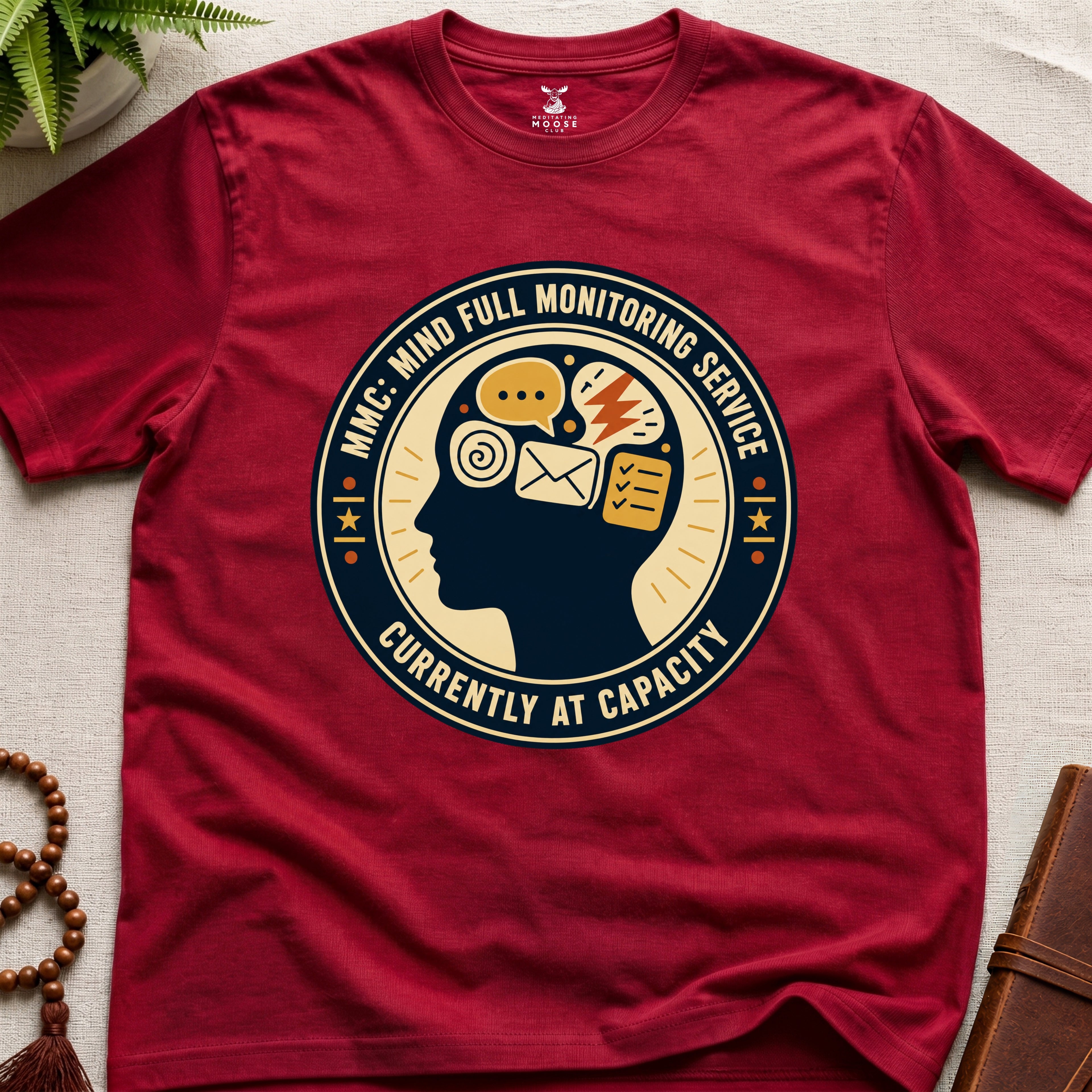 Red t-shirt with a badge graphic design featuring a head full of icons and text "Mind Full Monitoring Service" on a beige surface