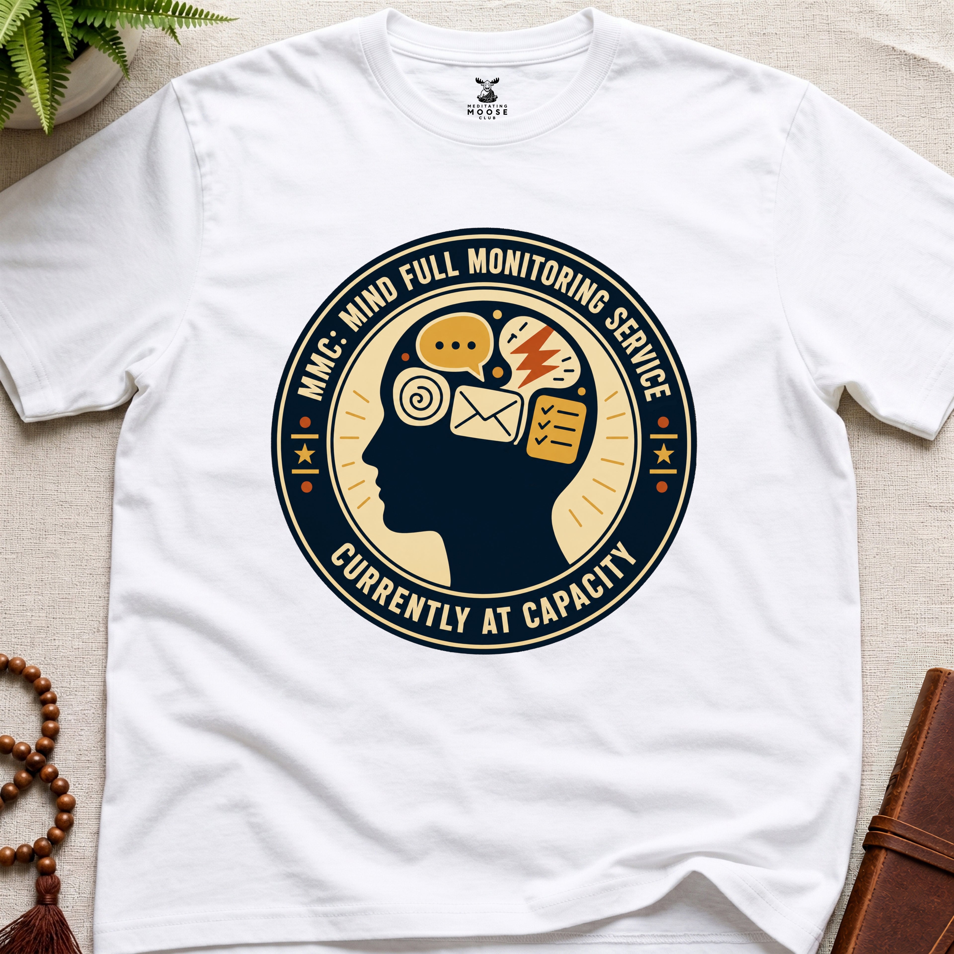 White t-shirt with a badge graphic design featuring a head full of icons and text "Mind Full Monitoring Service" on a beige surface