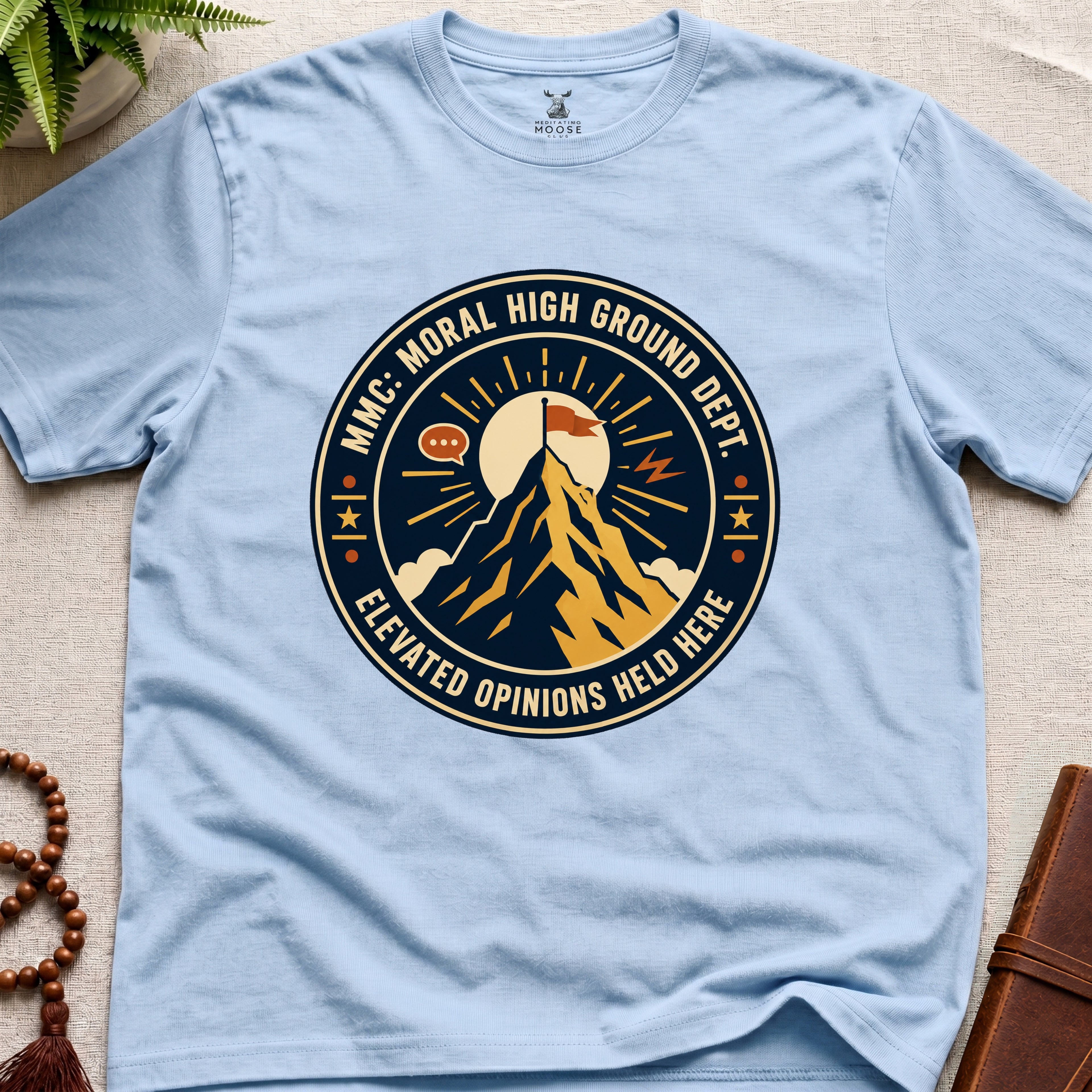 Light blue t-shirt badge graphic with a mountain and text "moral High Ground Dept." on a light surface.