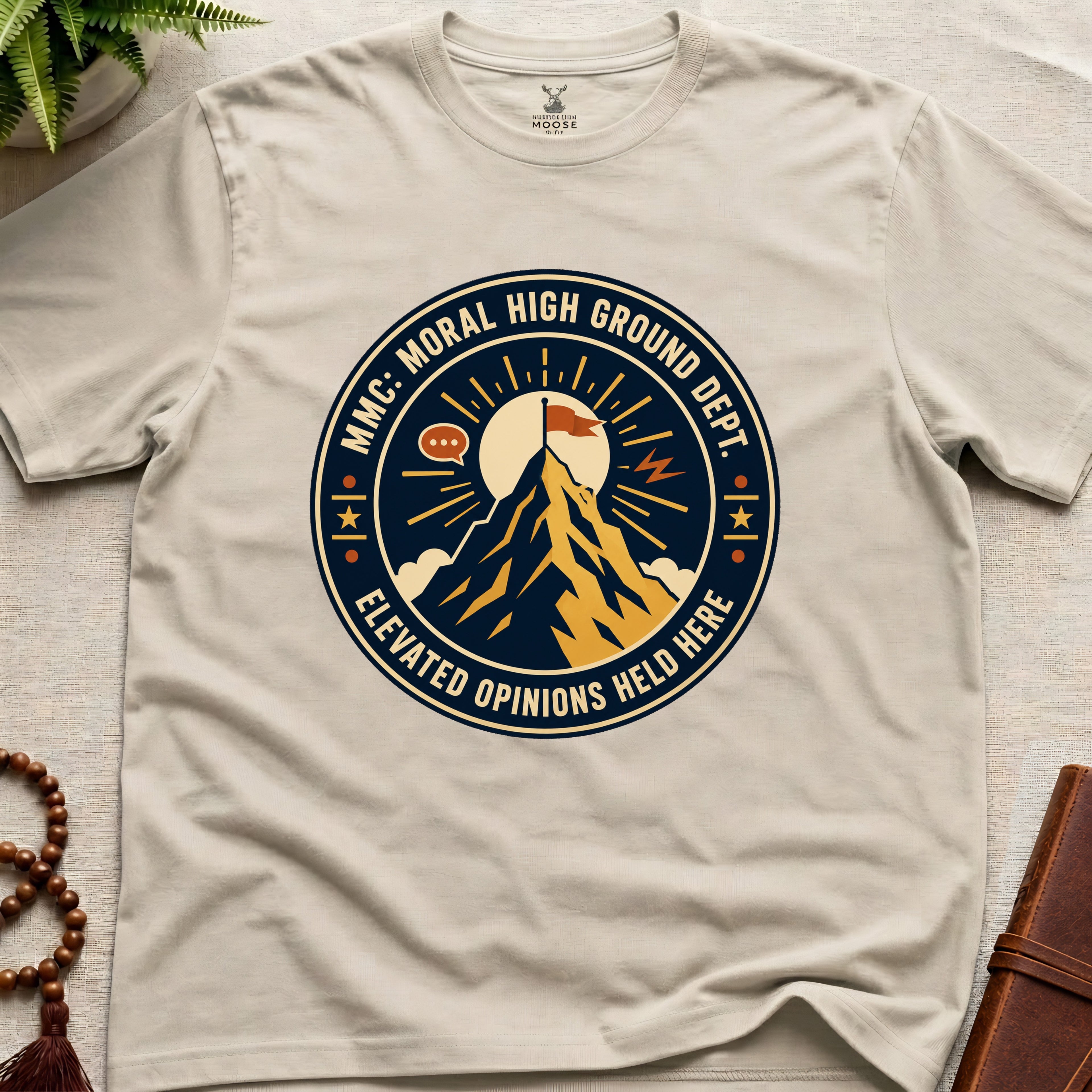 Beige t-shirt badge graphic with a mountain and text "moral High Ground Dept." on a light surface.