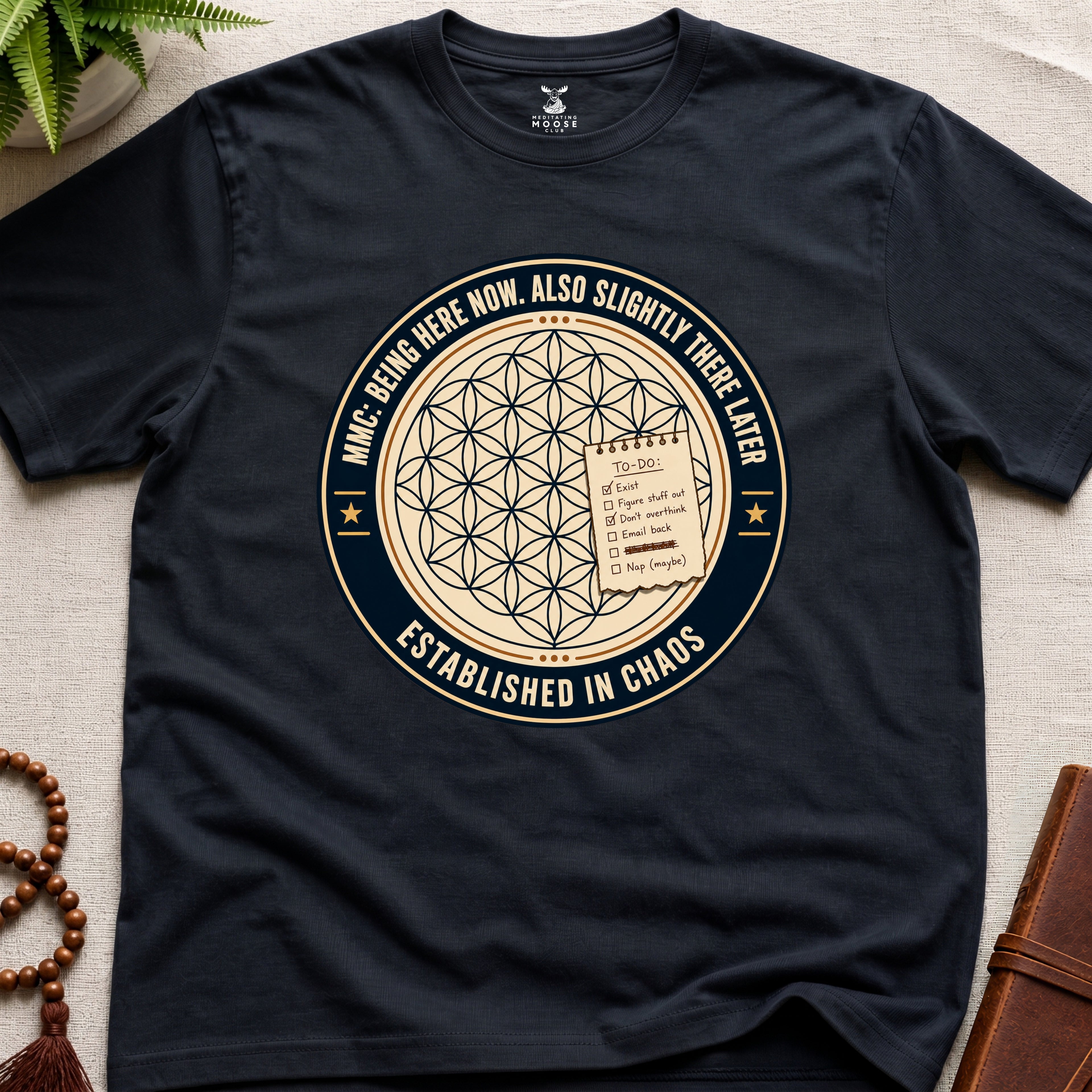 Black t-shirt with a circular badge design featuring text "established in chaos" and sacred geometry graphics on a light wooden surface.