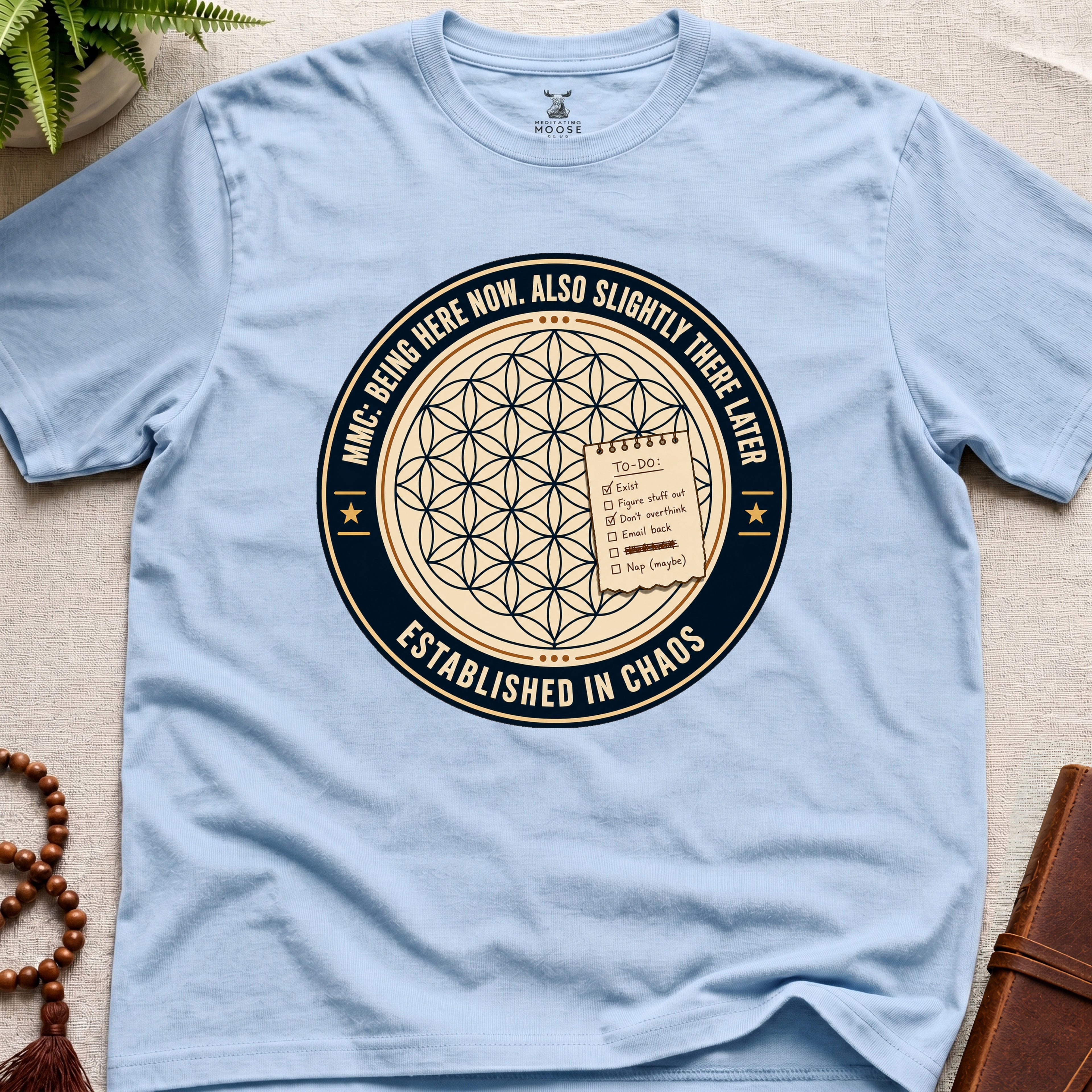 Light blue t-shirt with a circular badge design featuring text "established in chaos" and sacred geometry graphics on a light wooden surface.