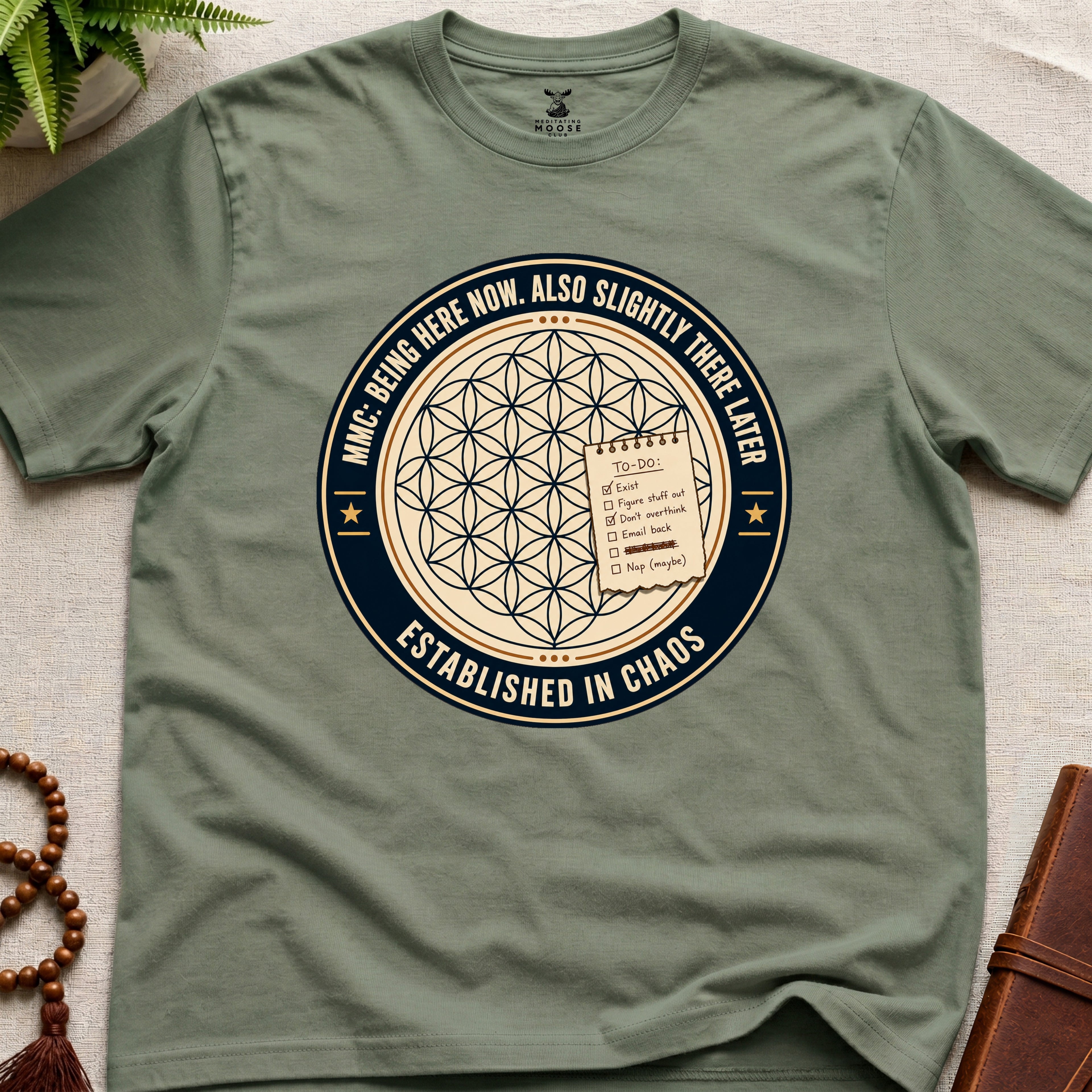 Military green t-shirt with a circular badge design featuring text "established in chaos" and sacred geometry graphics on a light wooden surface.
