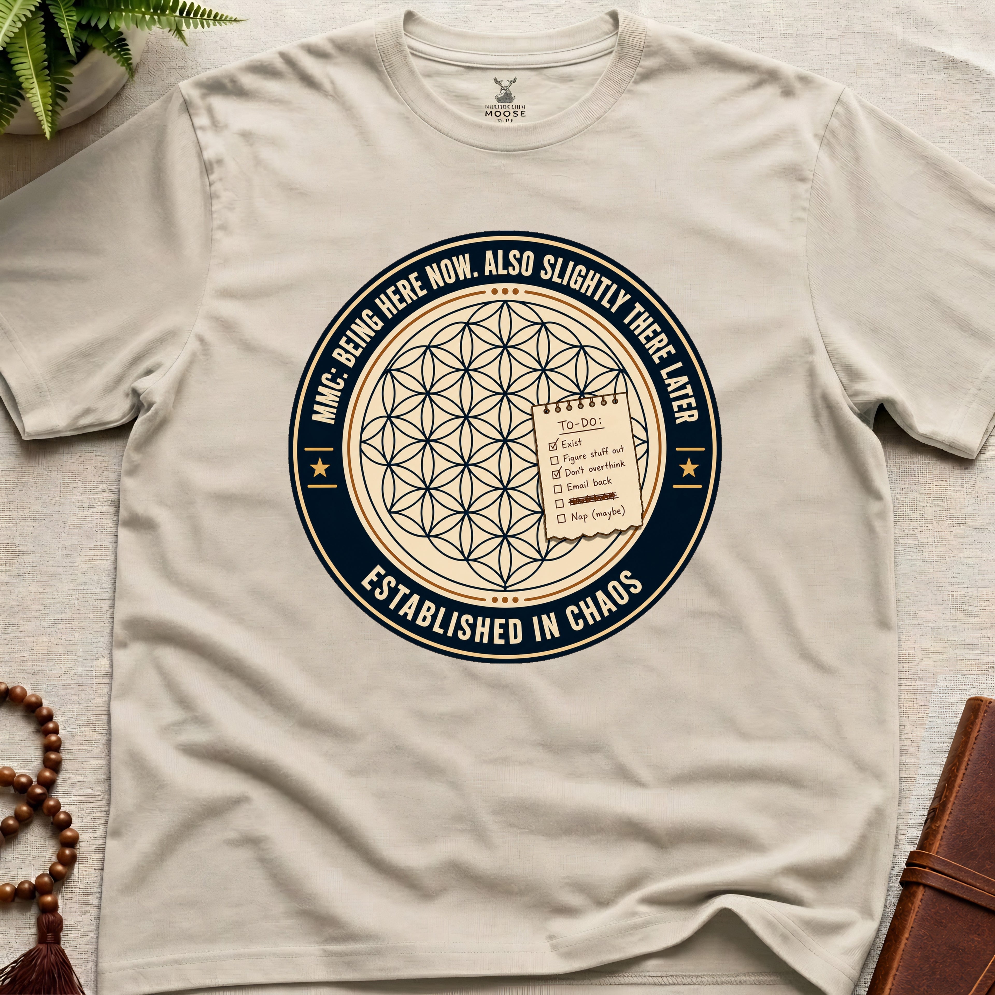 beige t-shirt with a circular badge design featuring text "established in chaos" and sacred geometry graphics on a light wooden surface.