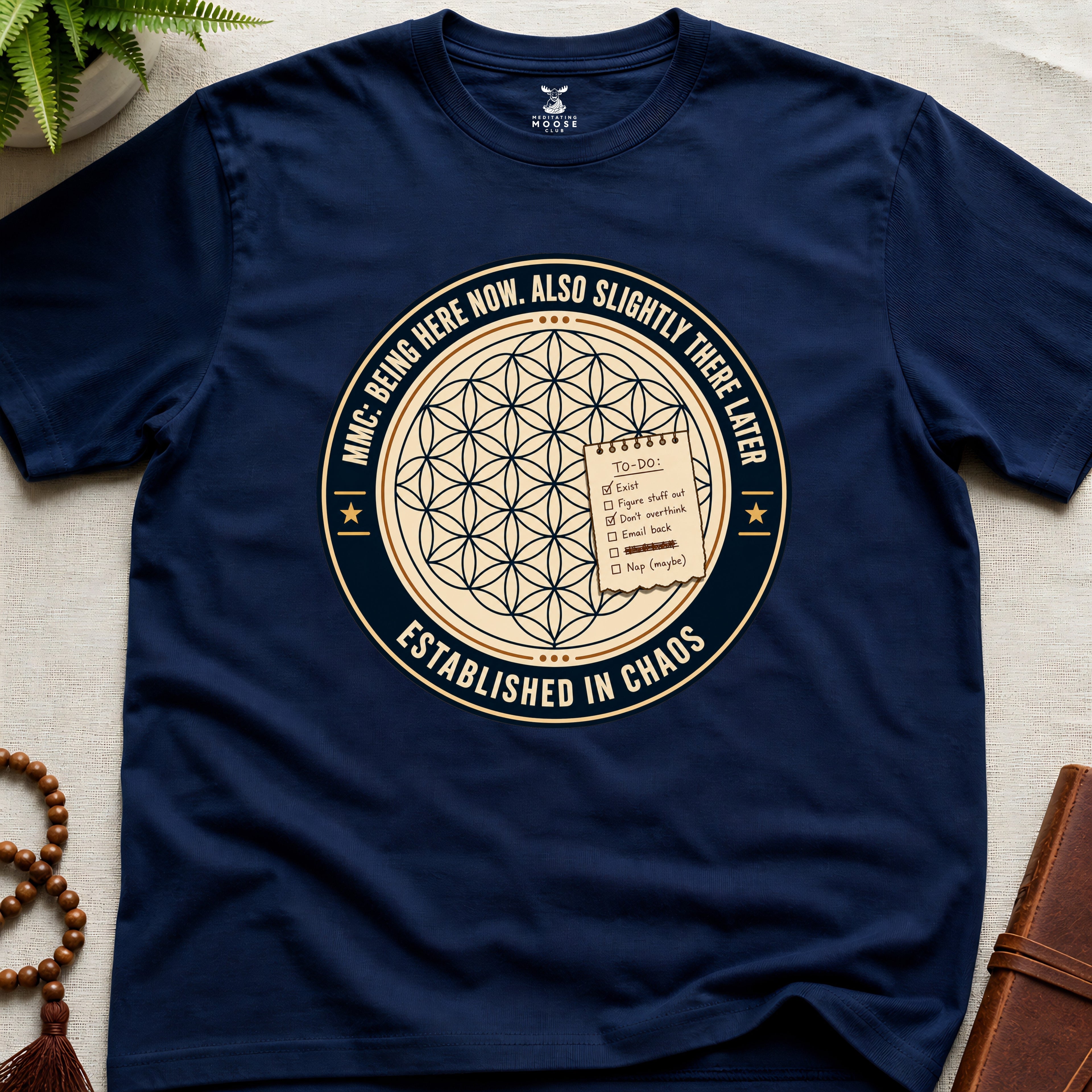 Navy t-shirt with a circular badge design featuring text "established in chaos" and sacred geometry graphics on a light wooden surface.