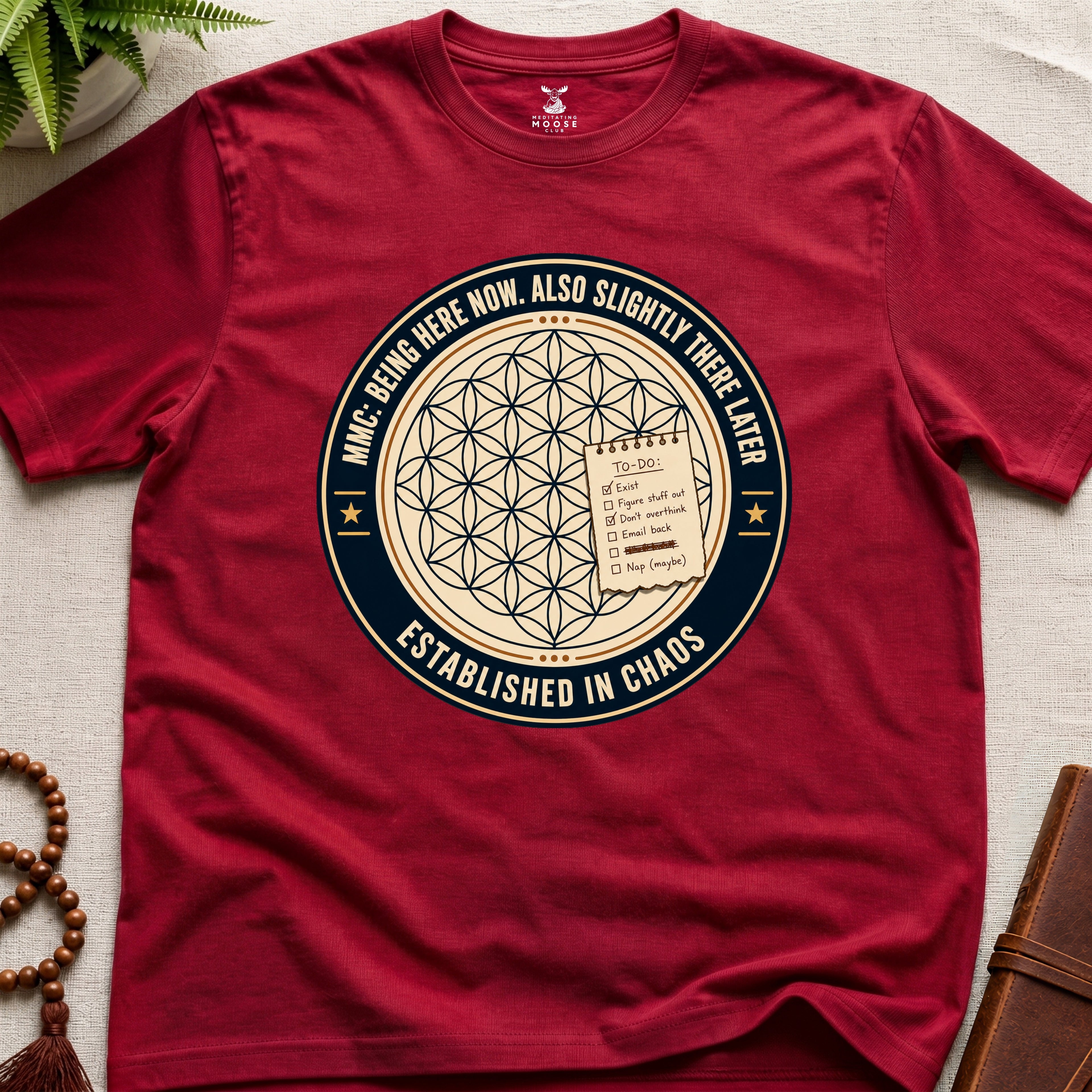 Red t-shirt with a circular badge design featuring text "established in chaos" and sacred geometry graphics on a light wooden surface.