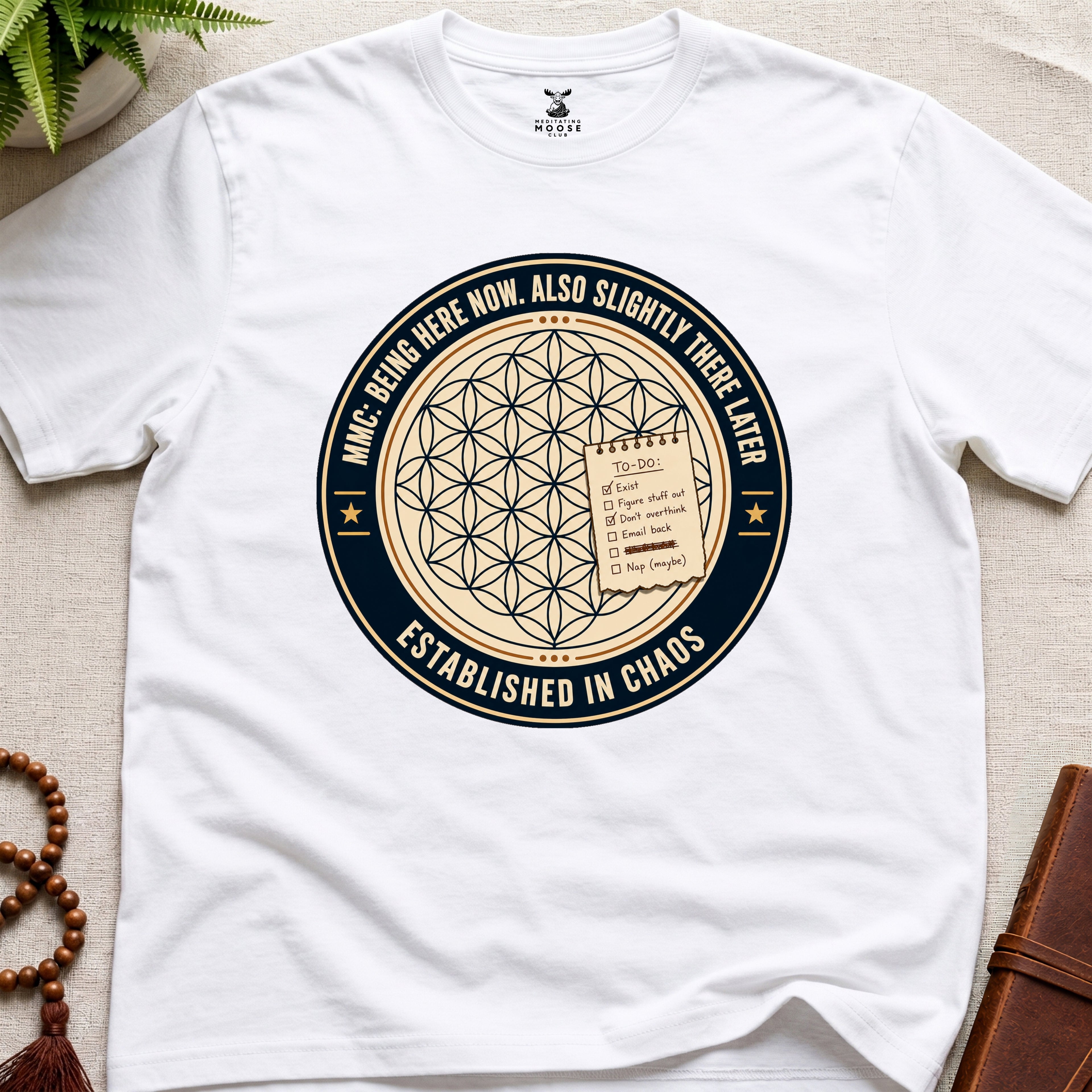 White t-shirt with a circular badge design featuring text "established in chaos" and sacred geometry graphics on a light wooden surface.