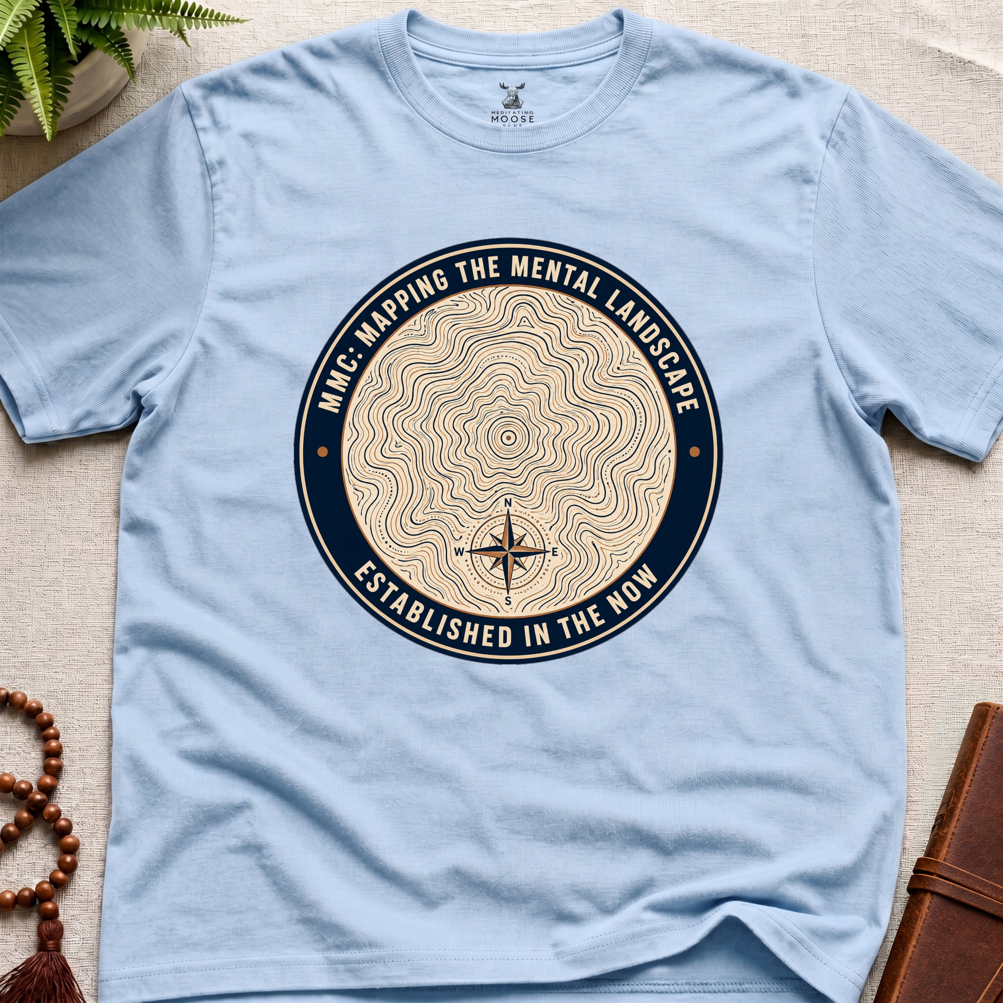 Light blue mindfulness t-shirt with topographic landscape badge design featuring compass