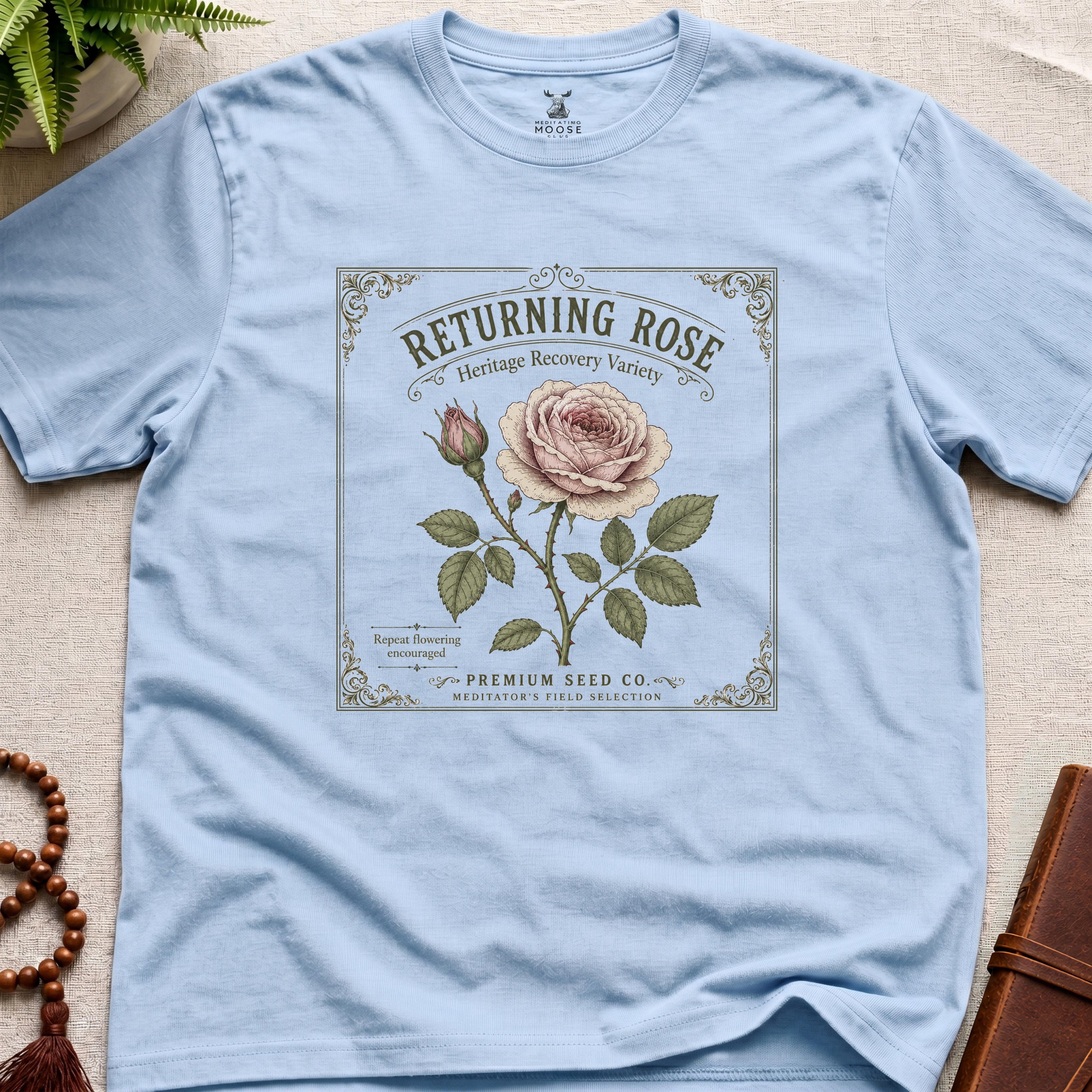 Light blue t-shirt with vintage see packet style, returning rose design on a beige surface.