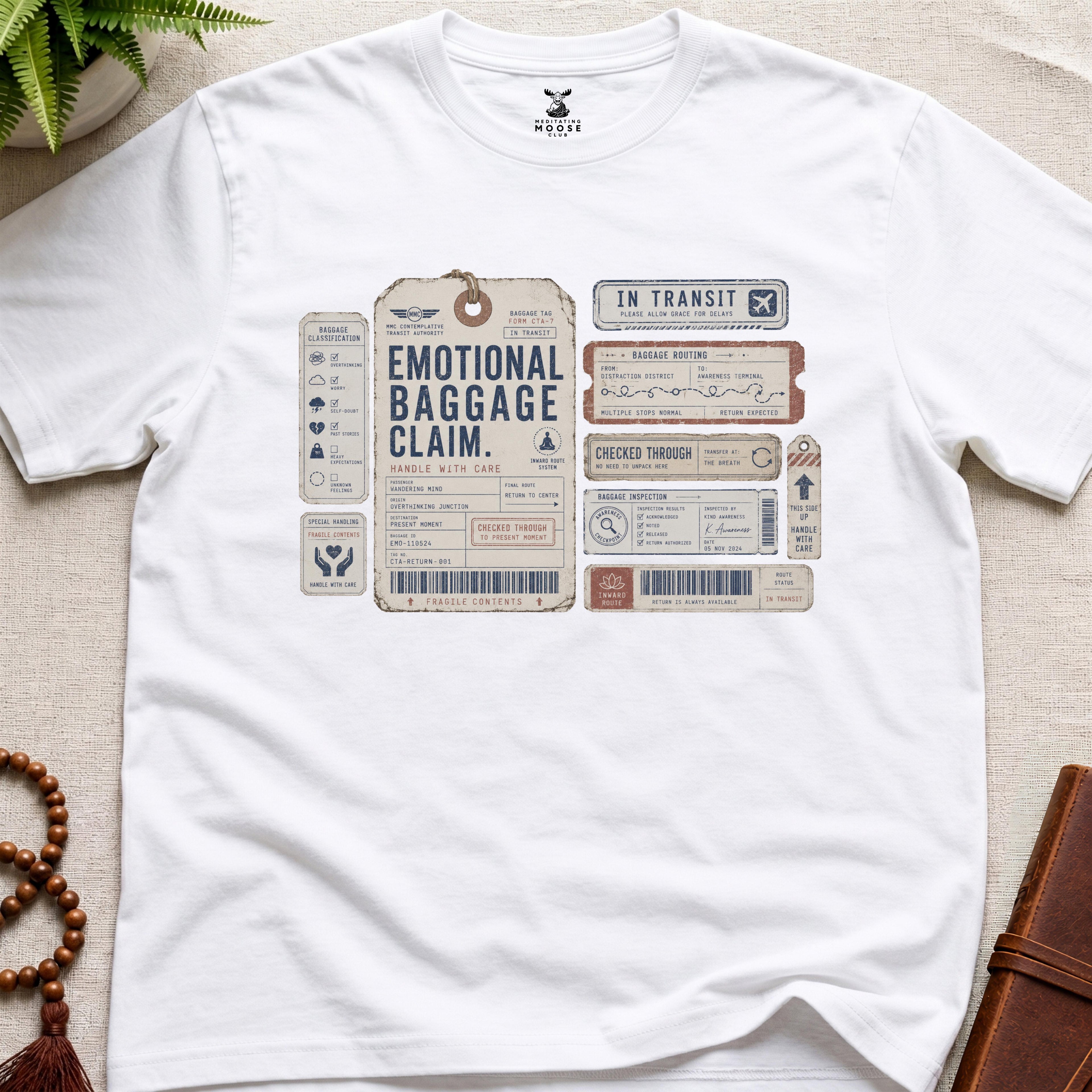 White vintage baggage-tag inspired mindfulness t-shirt graphic reading “Emotional Baggage Claim” with contemplative transit handling marks.