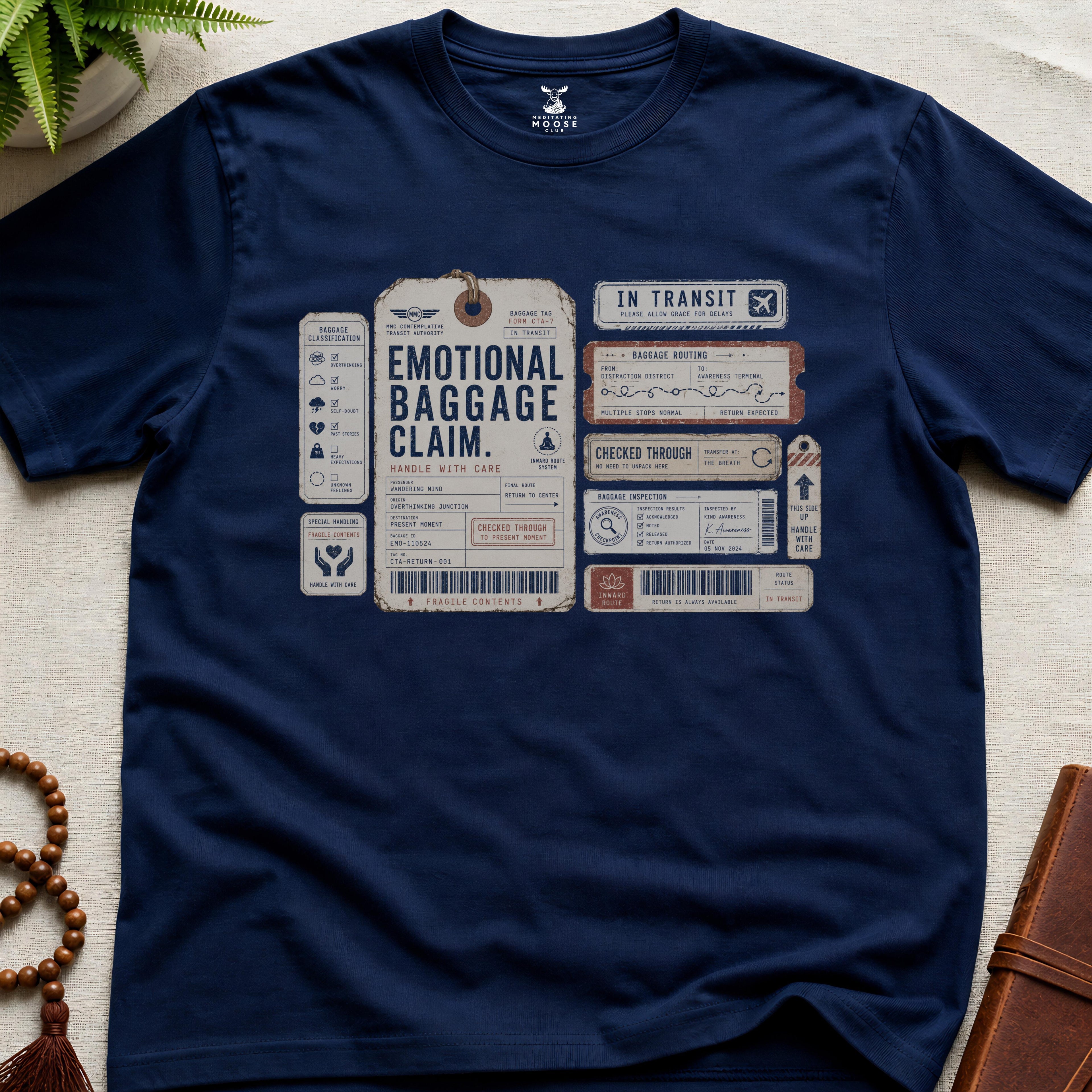 Navy vintage baggage-tag inspired mindfulness t-shirt graphic reading “Emotional Baggage Claim” with contemplative transit handling marks.