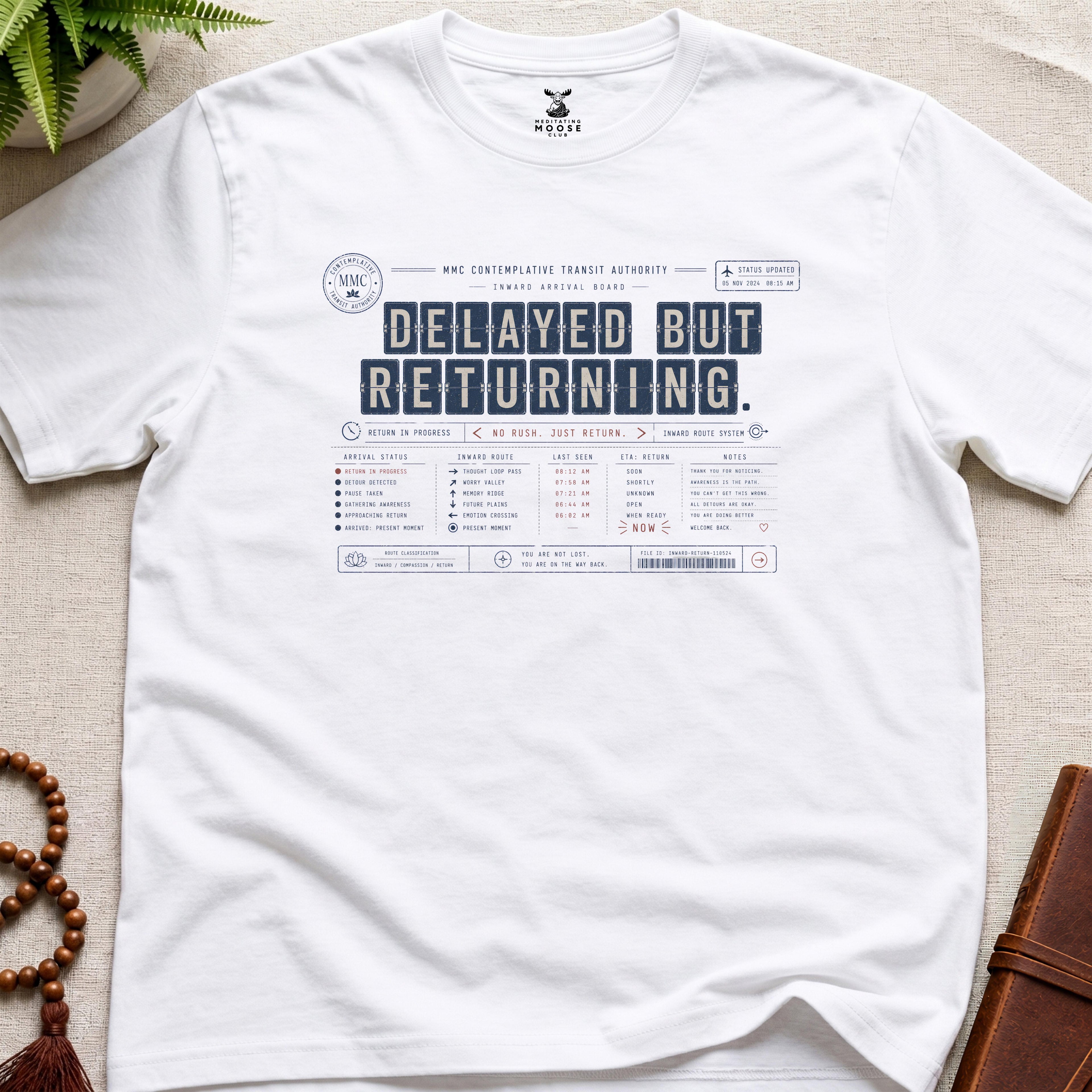 White vintage arrival-board inspired meditation t-shirt reading “Delayed But Returning” with contemplative transit typography.
