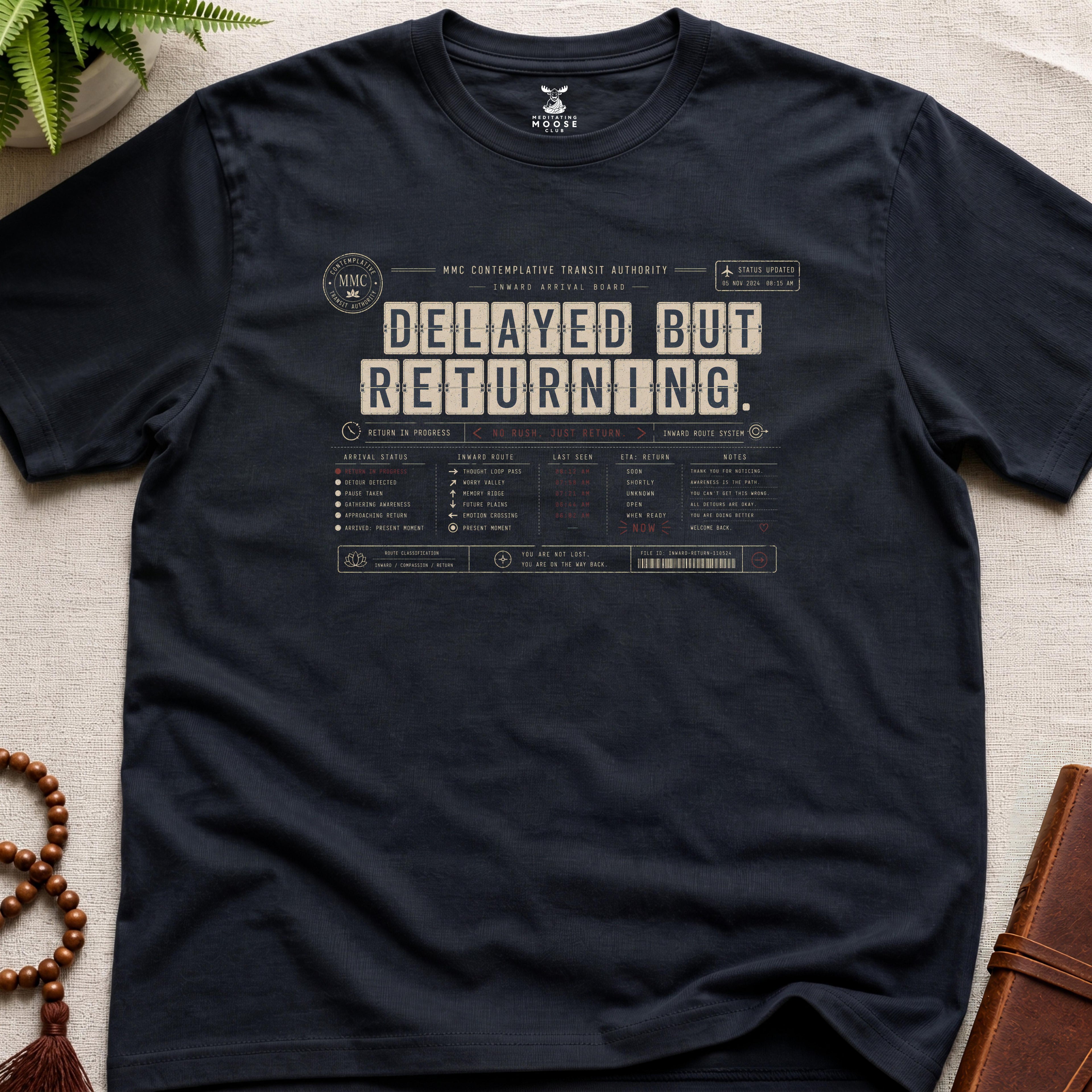 Black vintage arrival-board inspired meditation t-shirt reading “Delayed But Returning” with contemplative transit typography.