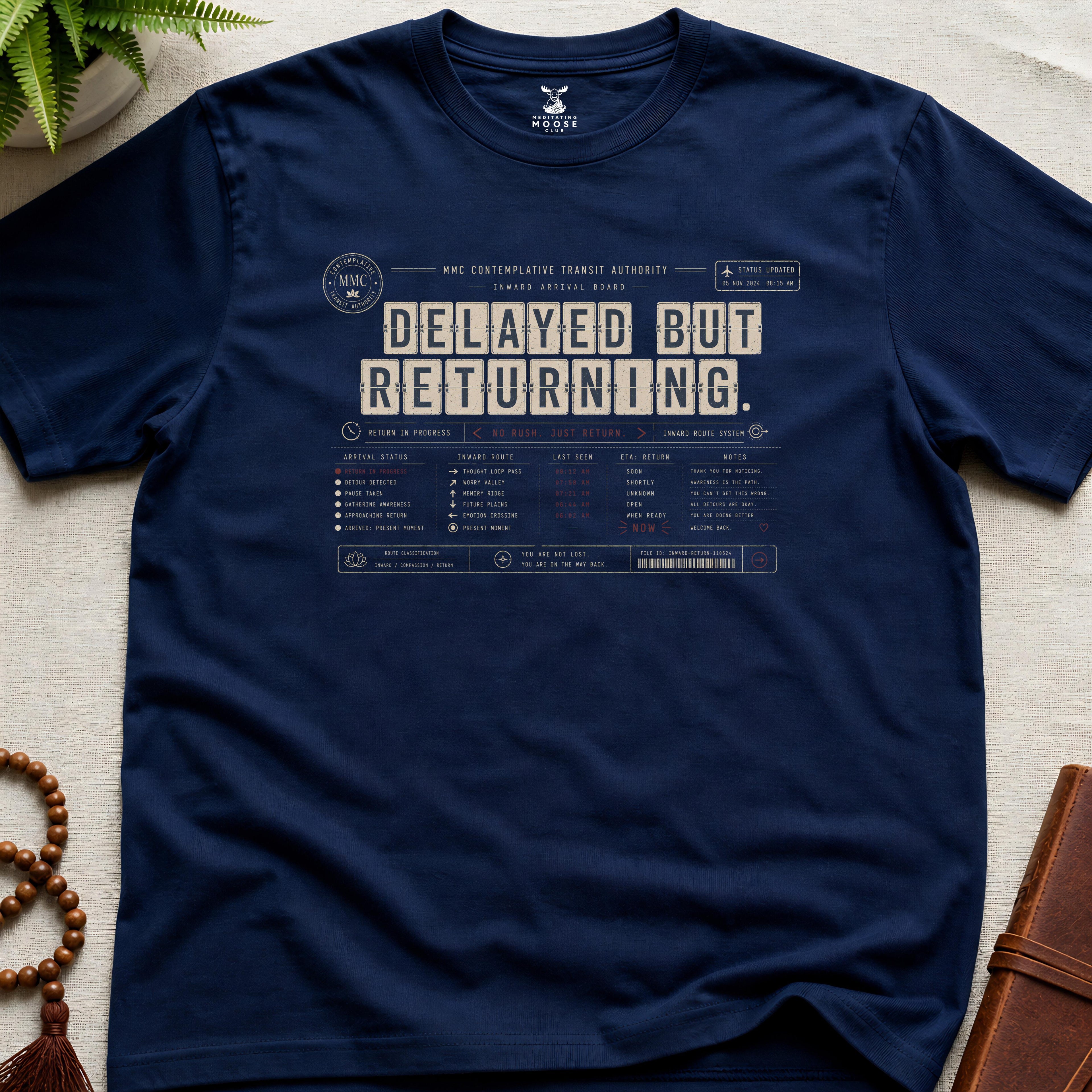 Navy vintage arrival-board inspired meditation t-shirt reading “Delayed But Returning” with contemplative transit typography.