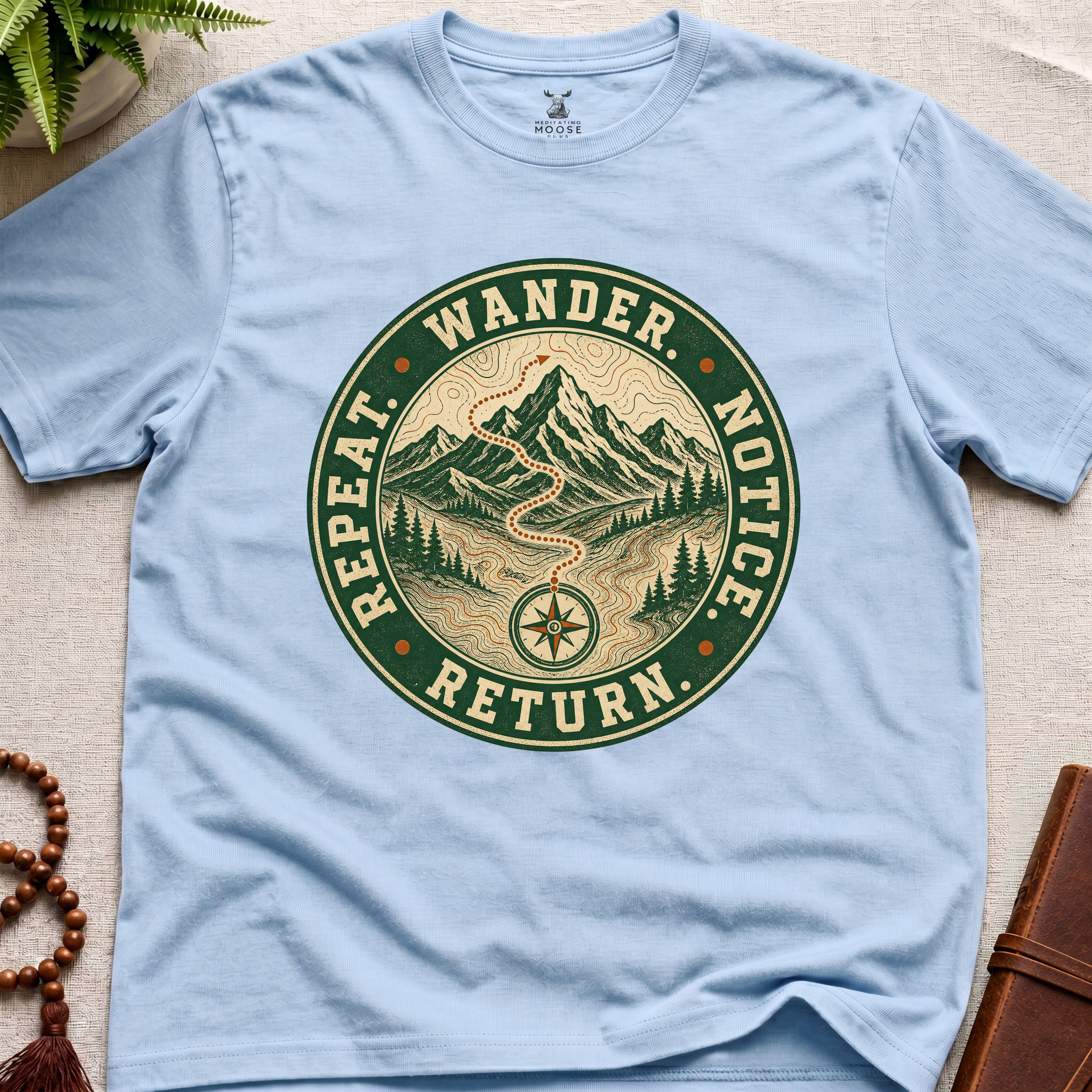 Light blue mindfulness t-shirt mockup featuring Wander Notice Return vintage mountain meditation badge graphic