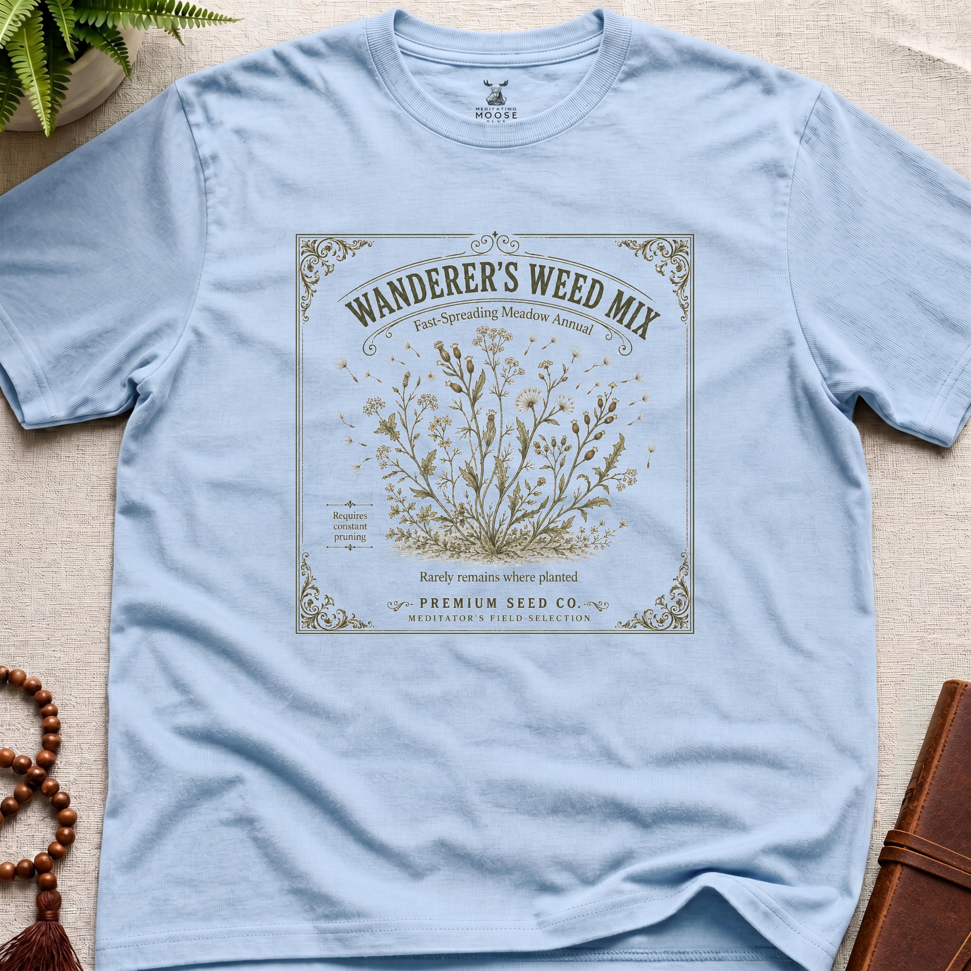 Light blue t-shirt with 'Wanderer's Weed Mix' seed packet design on a beige surface.